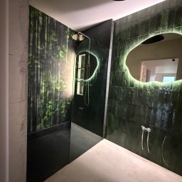 DAN-HOUSE SPÓŁKA Z OGRANICZONĄ ODPOWIEDZIALNOŚCIĄ - Nowoczesna łazienka z zielonymi płytkami i podświetlanym lustrem o nieregularnym kształcie. Widoczny prysznic i fragment ściany z motywem roślinnym.