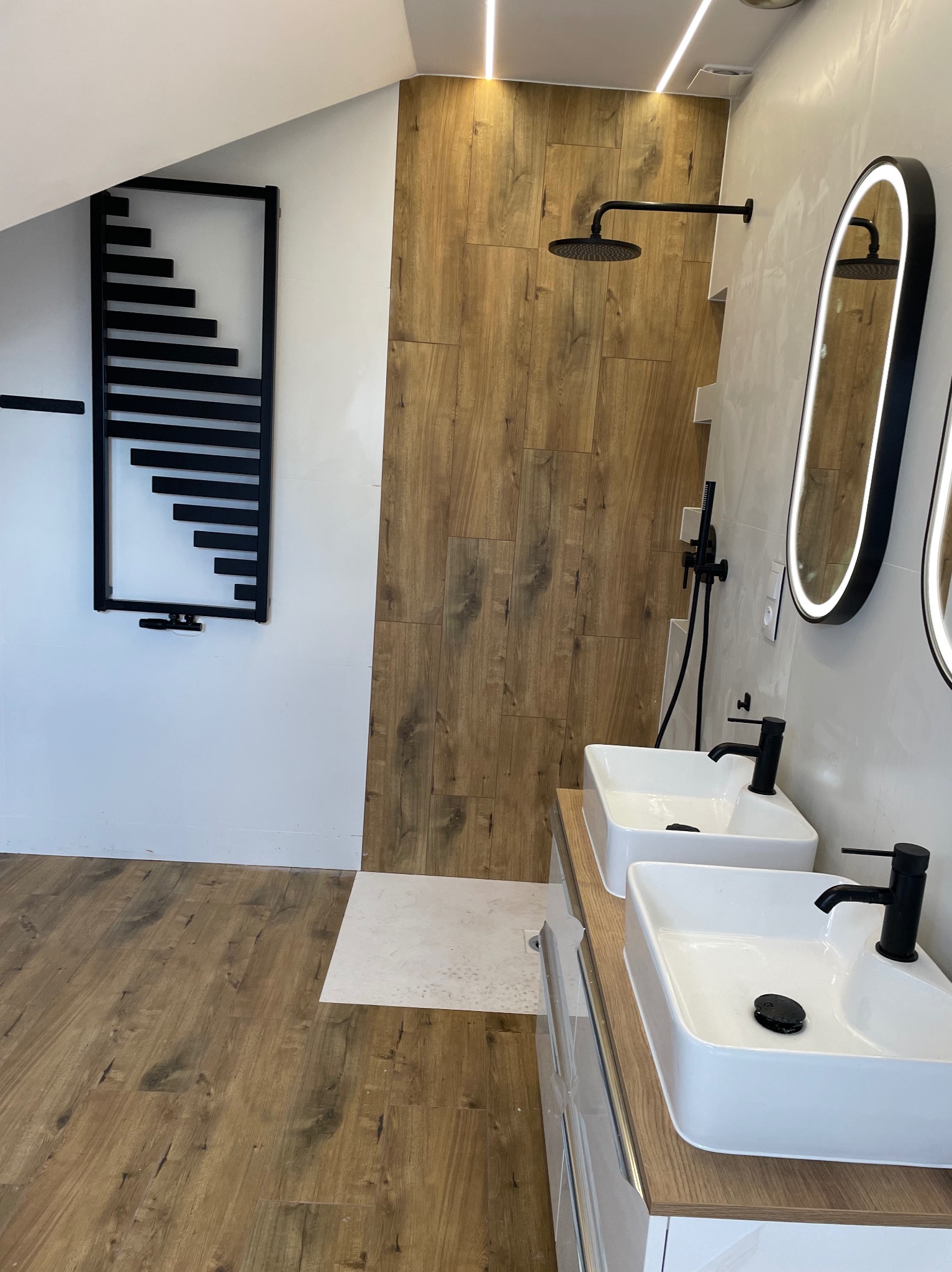 Nowoczesna łazienka z dwoma białymi umywalkami nablatowymi z czarnymi bateriami, czarnym prysznicem typu deszczownica, podświetlanymi lustrami owalnymi w czarnej ramie oraz czarnym grzejnikiem...