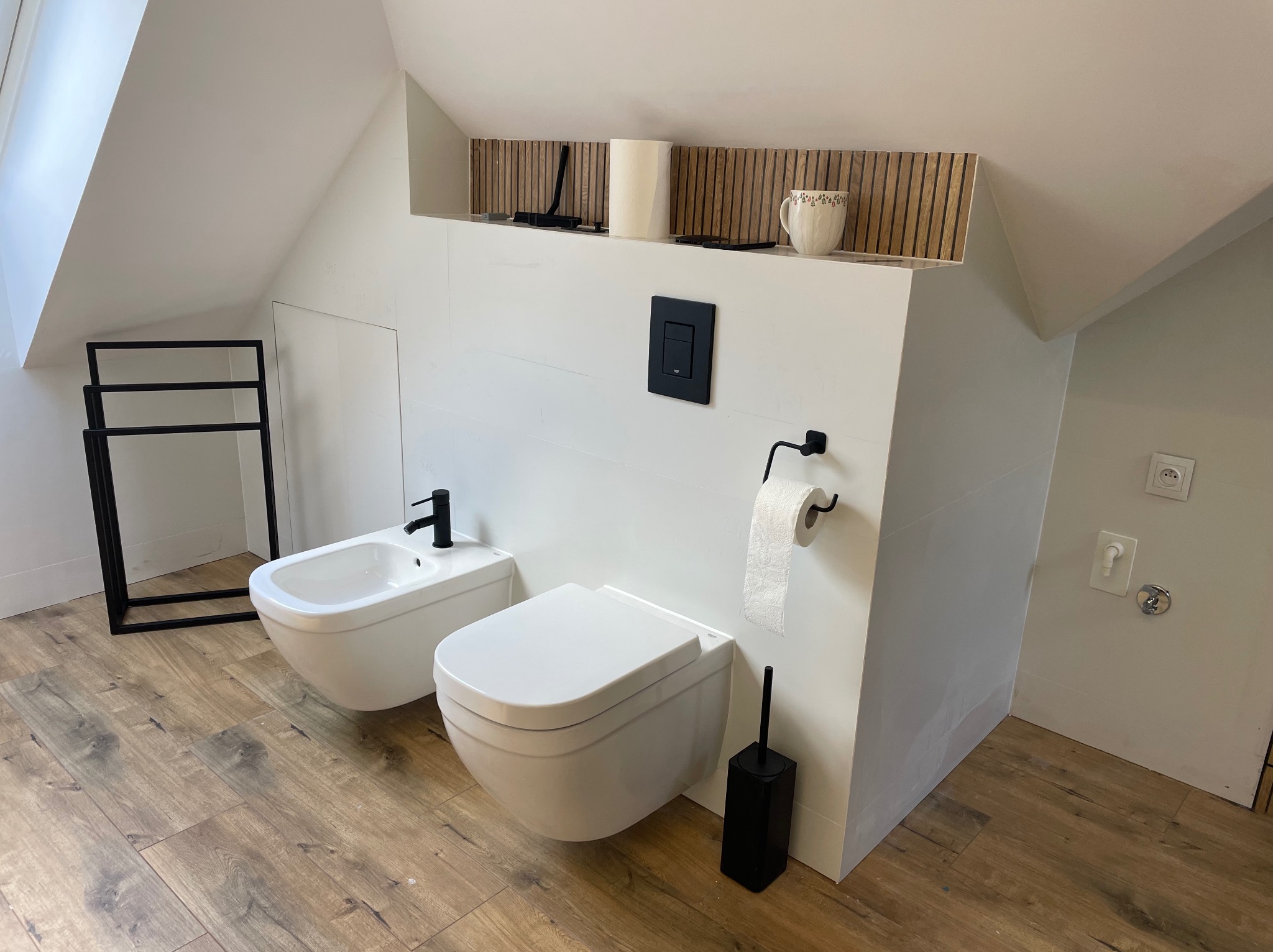 Nowoczesna łazienka na poddaszu z białymi płytkami, podwieszaną toaletą, bidetem i czarnymi akcentami: bateria, przycisk spłuczki, uchwyt na papier toaletowy, szczotka do WC i grzejnik drabinkowy.