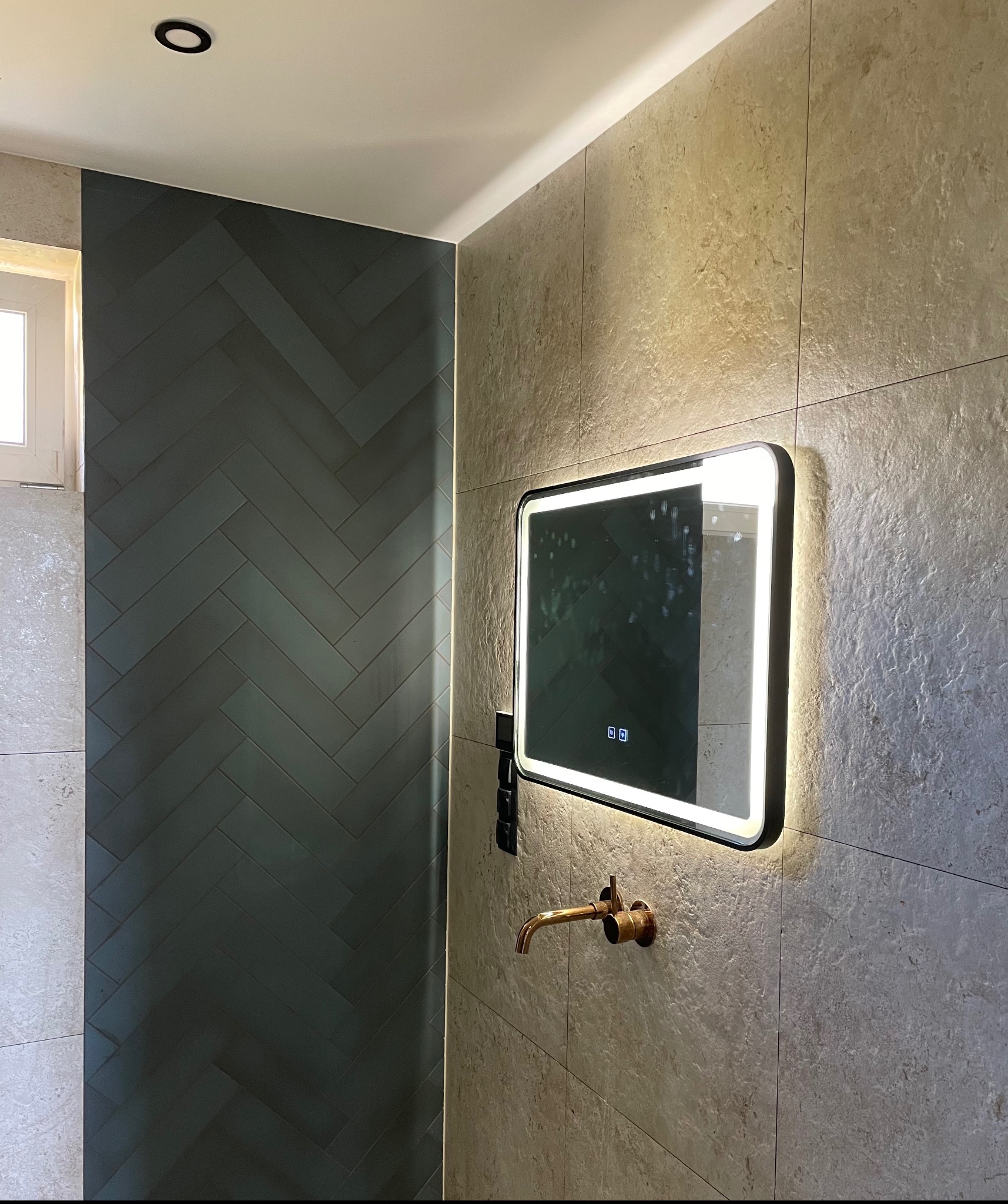 Nowoczesna łazienka z ciemnozielonymi płytkami ułożonymi w jodełkę, beżowymi płytkami imitującymi kamień, podświetlanym lustrem i miedzianą baterią.