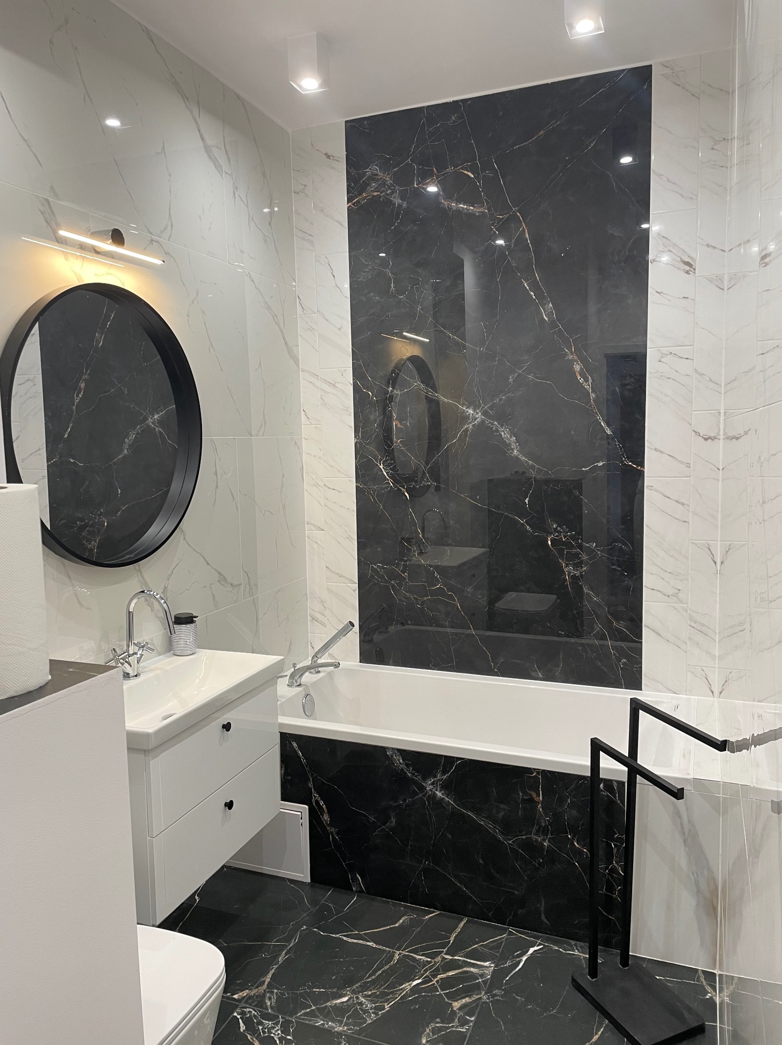 Nowoczesna łazienka we Wrocławiu z czarno-białymi marmurowymi płytkami, okrągłym lustrem z podświetleniem, białą umywalką z szafką i wanną z czarnym panelem.