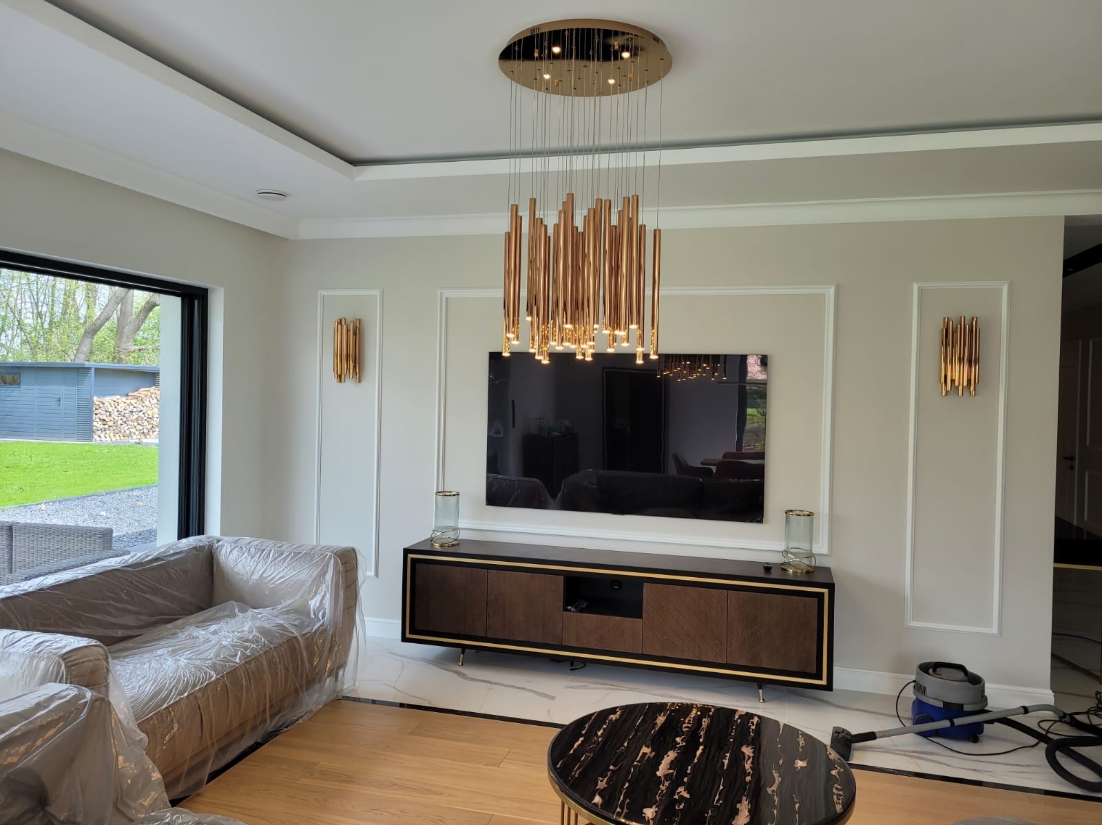 Salon po remoncie z nowoczesnym oświetleniem: wisząca lampa z złotych rurek, złote kinkiety, telewizor na ścianie, meble z drewnianymi frontami, marmurowa podłoga, sofa zabezpieczona folią...