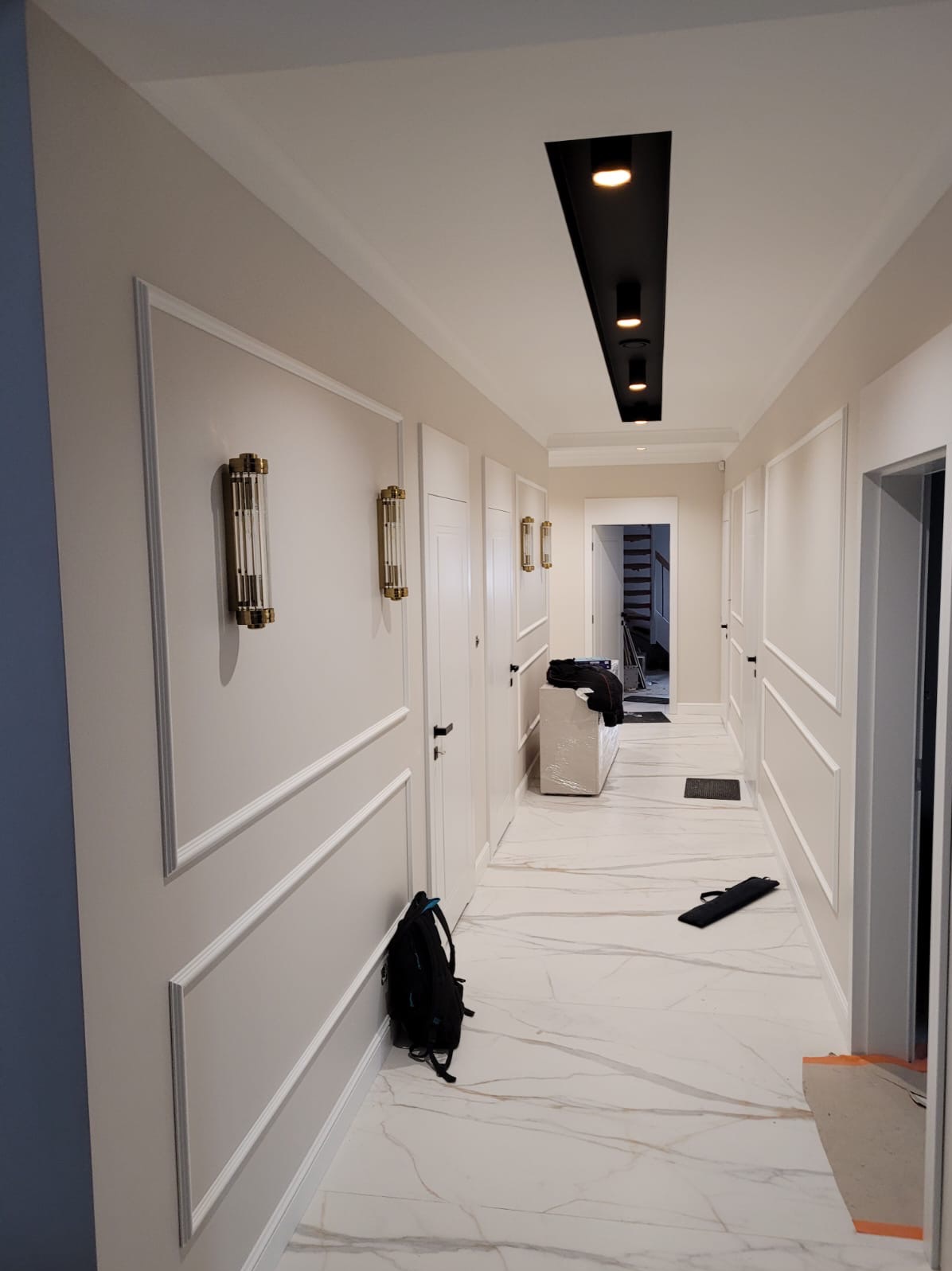 Nowoczesny korytarz po remoncie z białymi ścianami zdobionymi sztukaterią, marmurową podłogą, czarnym oświetleniem sufitowym i złotymi lampami ściennymi, w tle widoczne schody.
