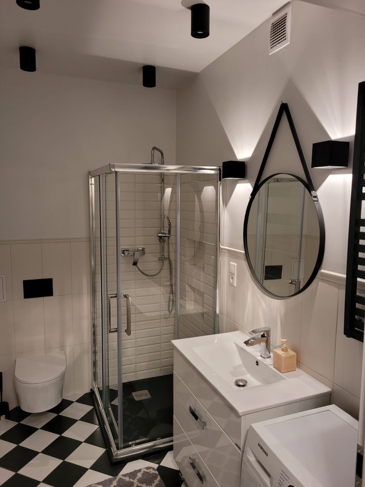 Nowoczesna łazienka z kabiną prysznicową, umywalką z szafką i okrągłym lustrem zawieszonym na czarnym pasku, czarno-biała szachownica na podłodze.