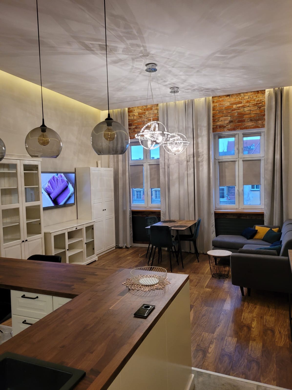 Przestronne wnętrze mieszkania po remoncie: drewniana podłoga, ściana z cegły, szare zasłony, szara sofa z kolorowymi poduszkami, stół z krzesłami, białe meble i nowoczesne lampy wiszące.