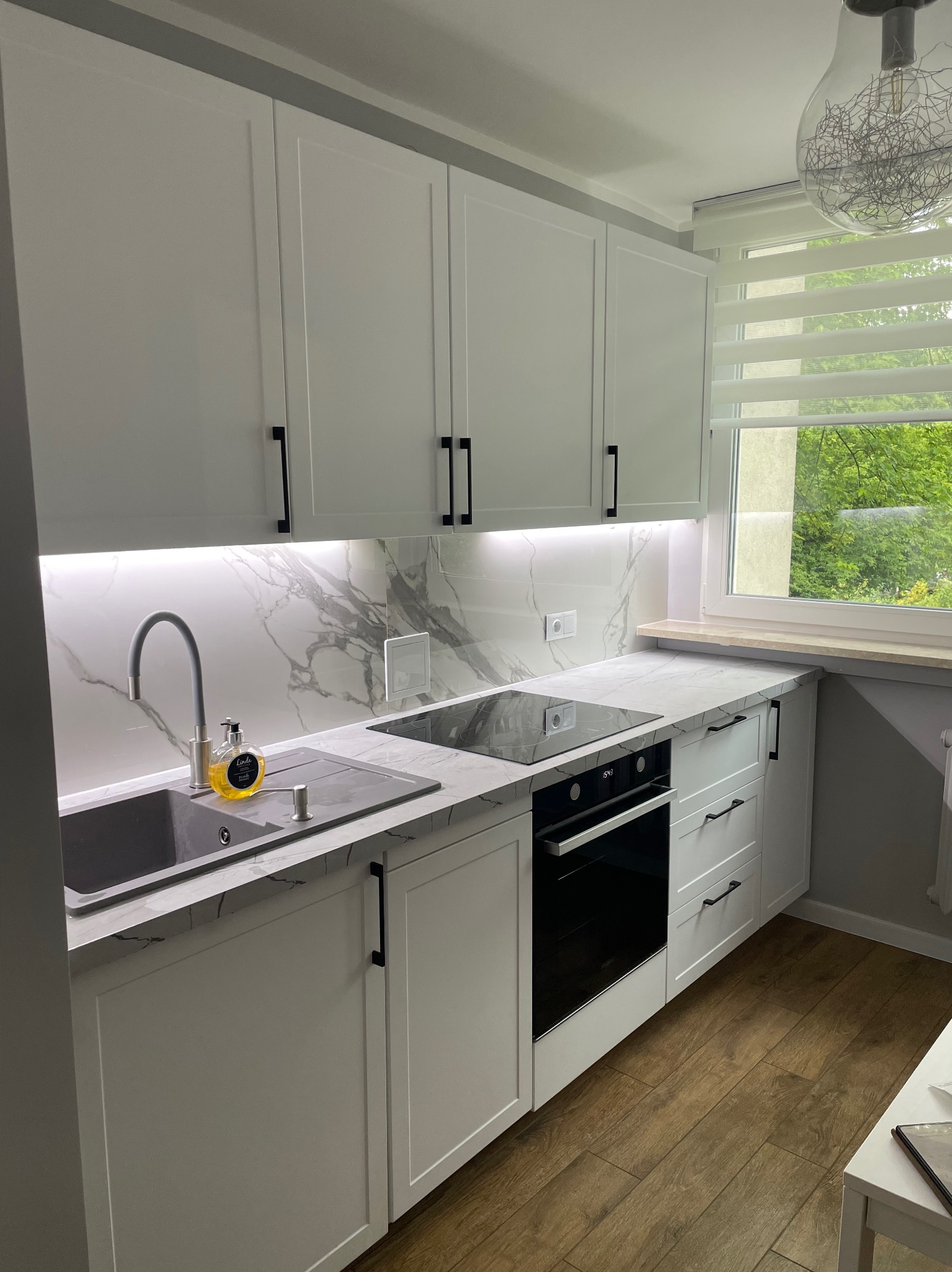 Nowoczesna, biała kuchnia z marmurowym blatem i backsplashiem, czarnymi uchwytami i drewnianą podłogą