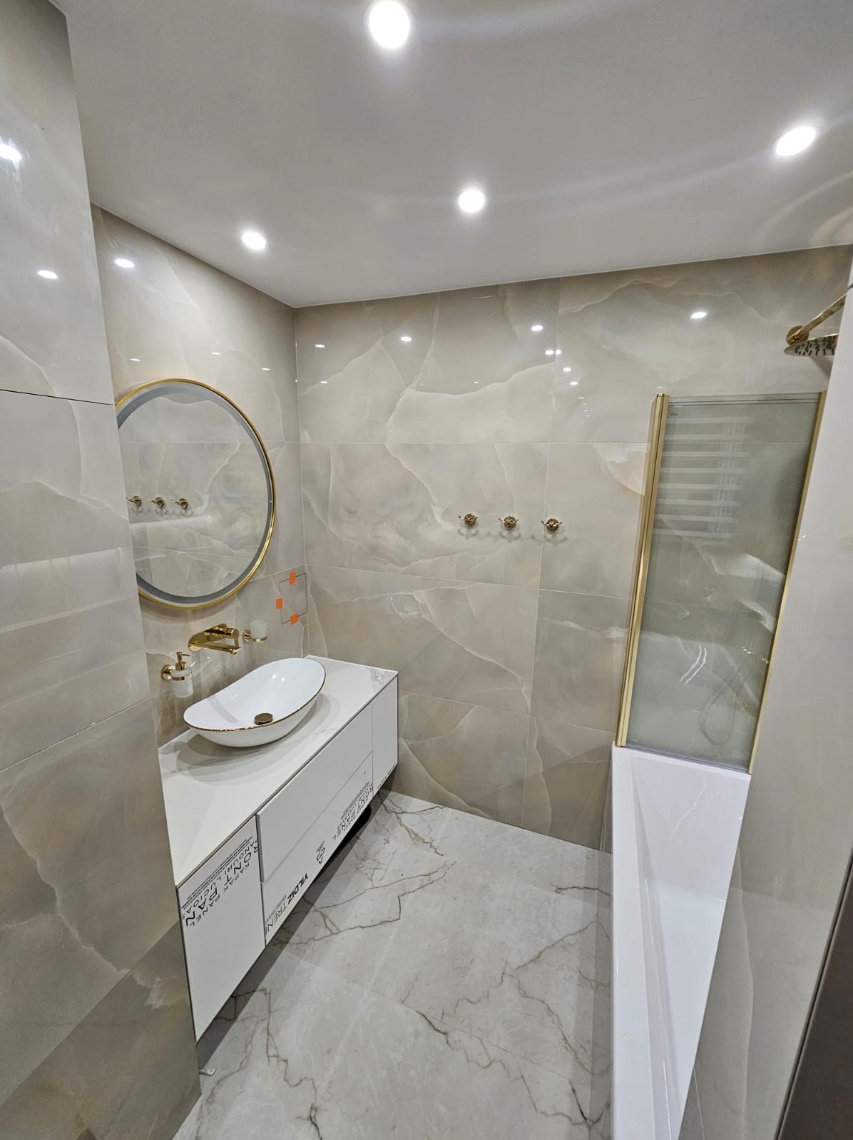 Elegancka łazienka z beżowymi płytkami imitującymi marmur, owalną umywalką na białej szafce, okrągłym lustrem w złotej ramie i kabiną prysznicową z elementami w kolorze złota.