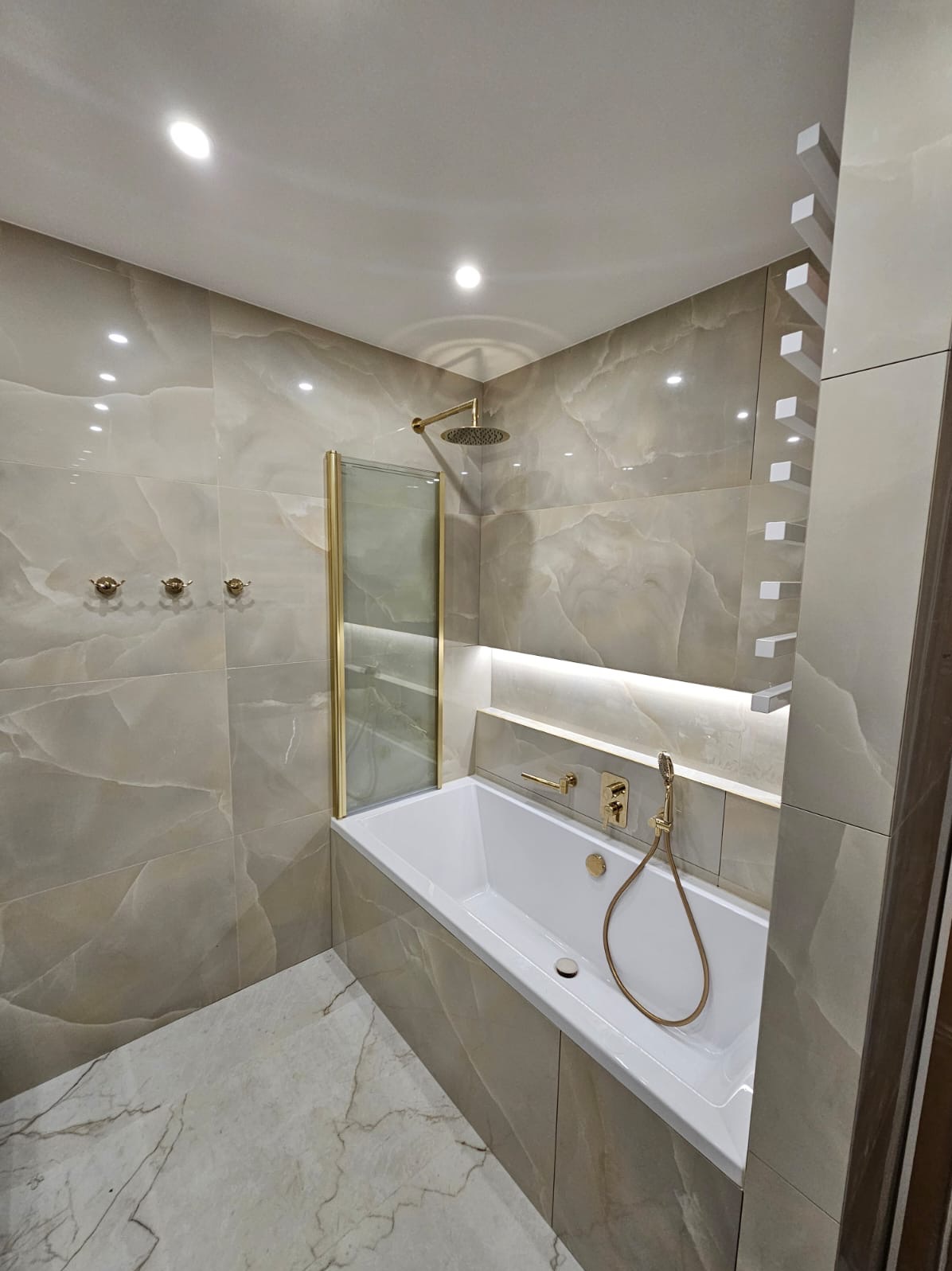Wykończona łazienka z wanną obudowaną płytkami imitującymi marmur, złotą armaturą i deszczownicą za szklanym panelem.