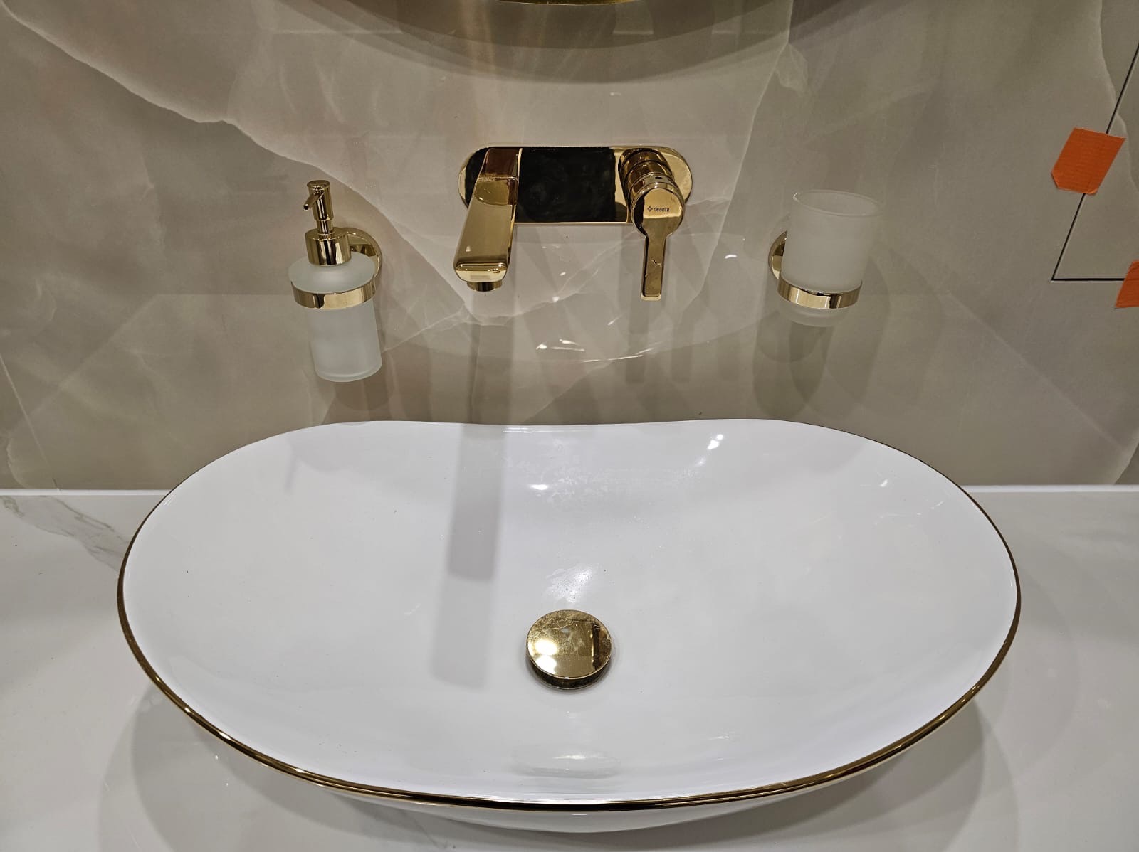 Biała, owalna umywalka z cienkim złotym obramowaniem i złotym odpływem, zamontowana na blacie; bateria ścienna w kolorze złotym, dozownik mydła i uchwyt na kubek w tym samym kolorze, marmurowe...