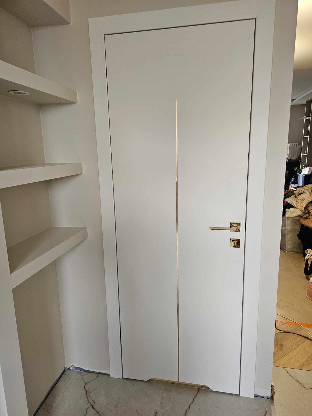 Białe, nowoczesne drzwi wewnętrzne z pionowym, złotym akcentem i złotą klamką, widoczne na tle białych półek wnękowych.