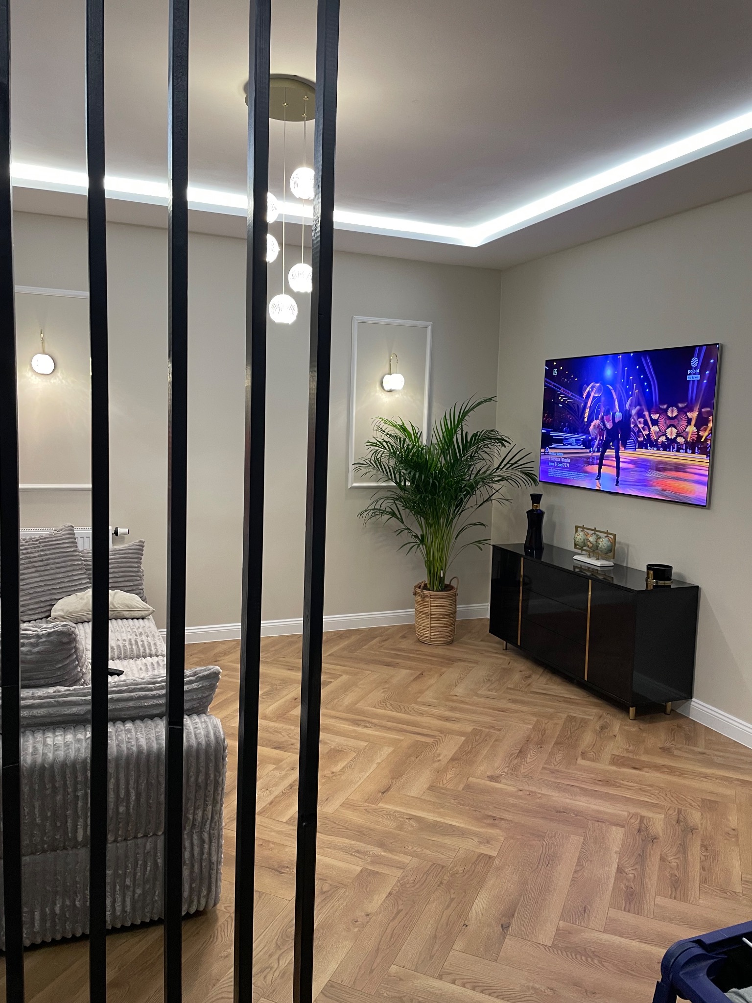 Salon z parkietem ułożonym w jodełkę, szarą sofą widoczną przez czarne, pionowe listwy, czarną komodą i rośliną doniczkową pod telewizorem.