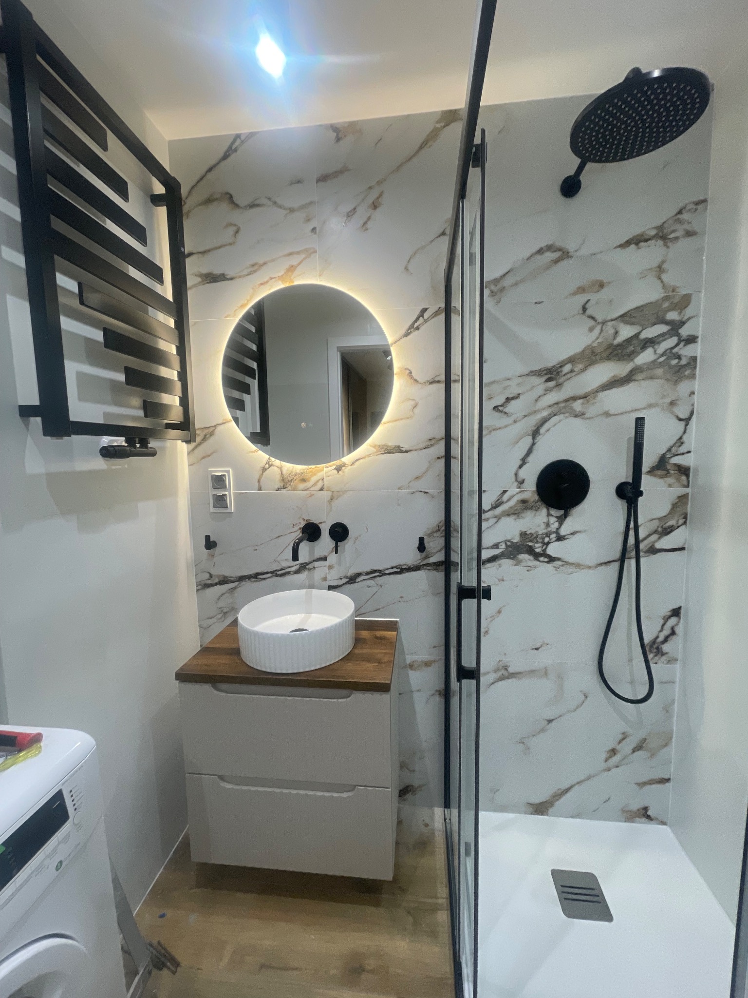 Nowoczesna łazienka z kabiną prysznicową typu walk-in, umywalką nablatową i okrągłym lustrem z podświetleniem LED. Marmurowe płytki na ścianach i czarne akcenty armatury.