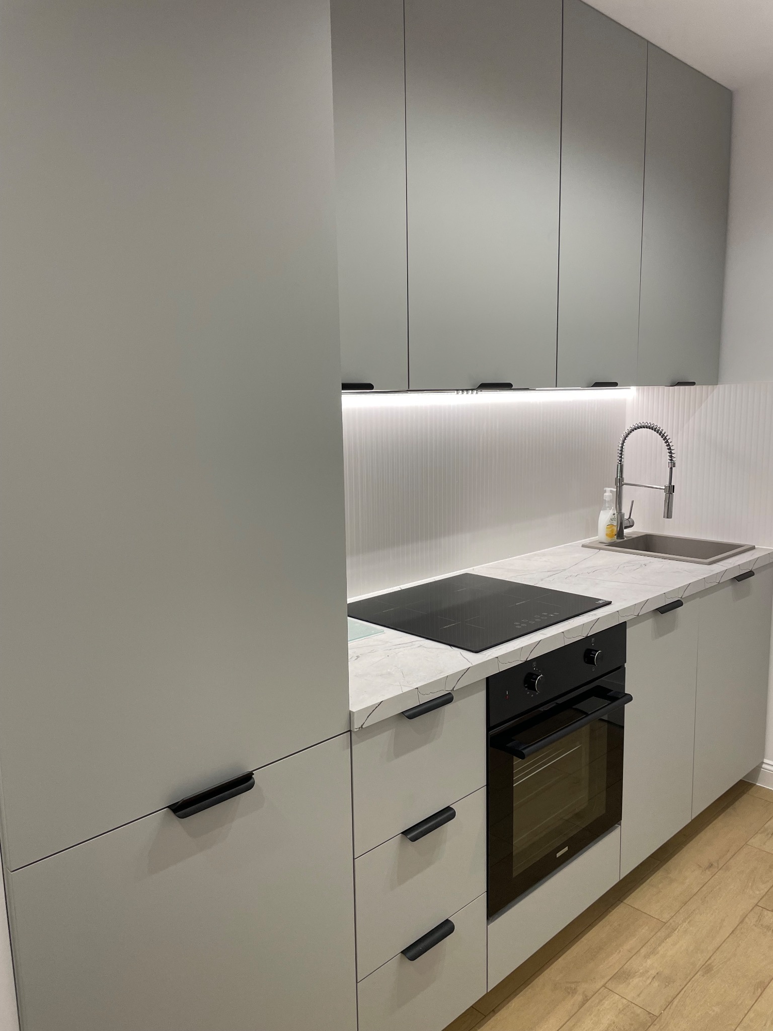 Nowoczesna, szara zabudowa kuchenna z marmurowym blatem, czarną płytą indukcyjną i piekarnikiem. Ujęcie z bliska, widoczne detale wykończenia i armatura.