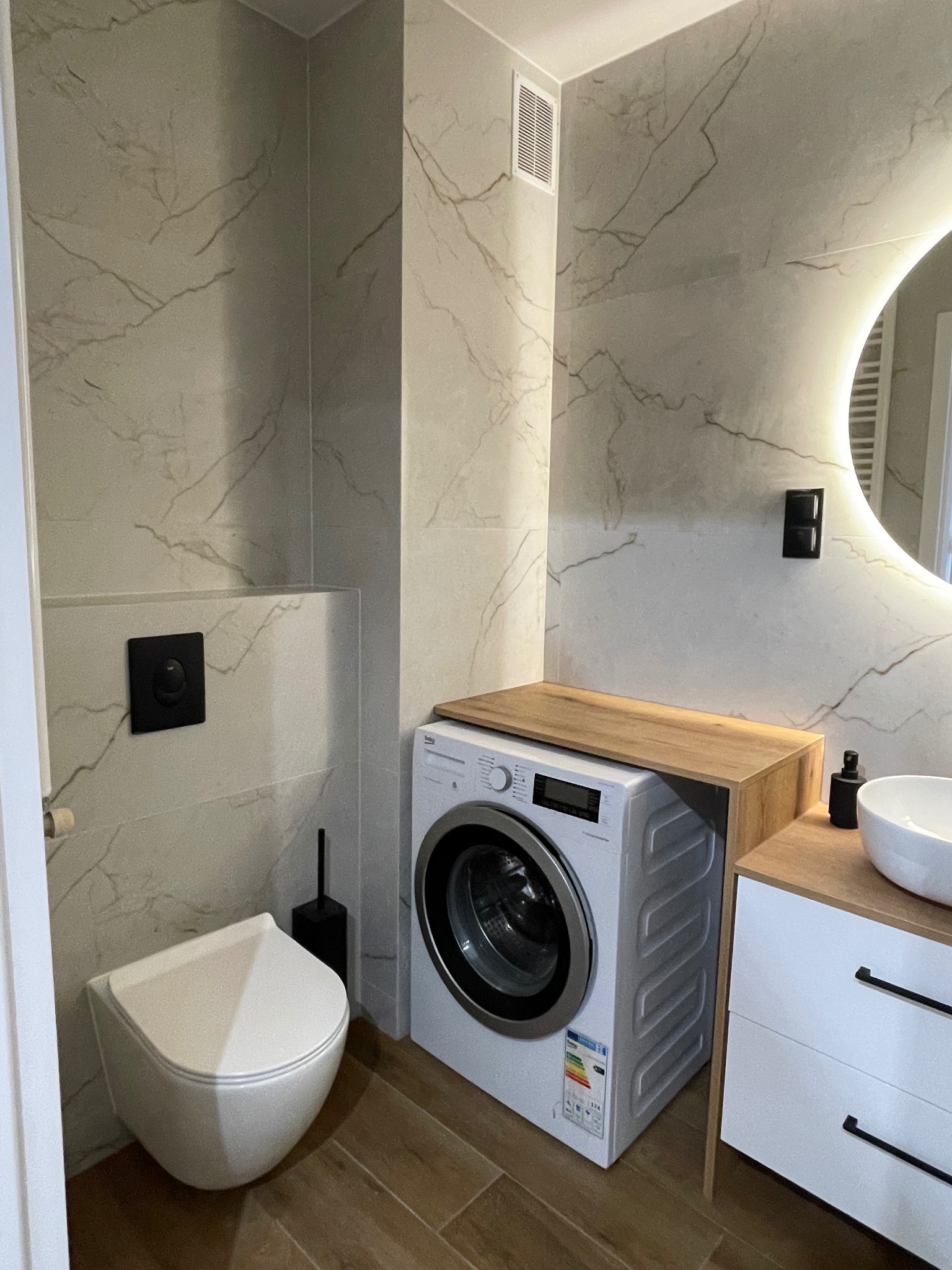 Nowoczesna łazienka z płytkami imitującymi marmur, wiszącą toaletą i pralką zabudowaną drewnianym blatem. Okrągłe lustro z podświetleniem, drewniana podłoga.