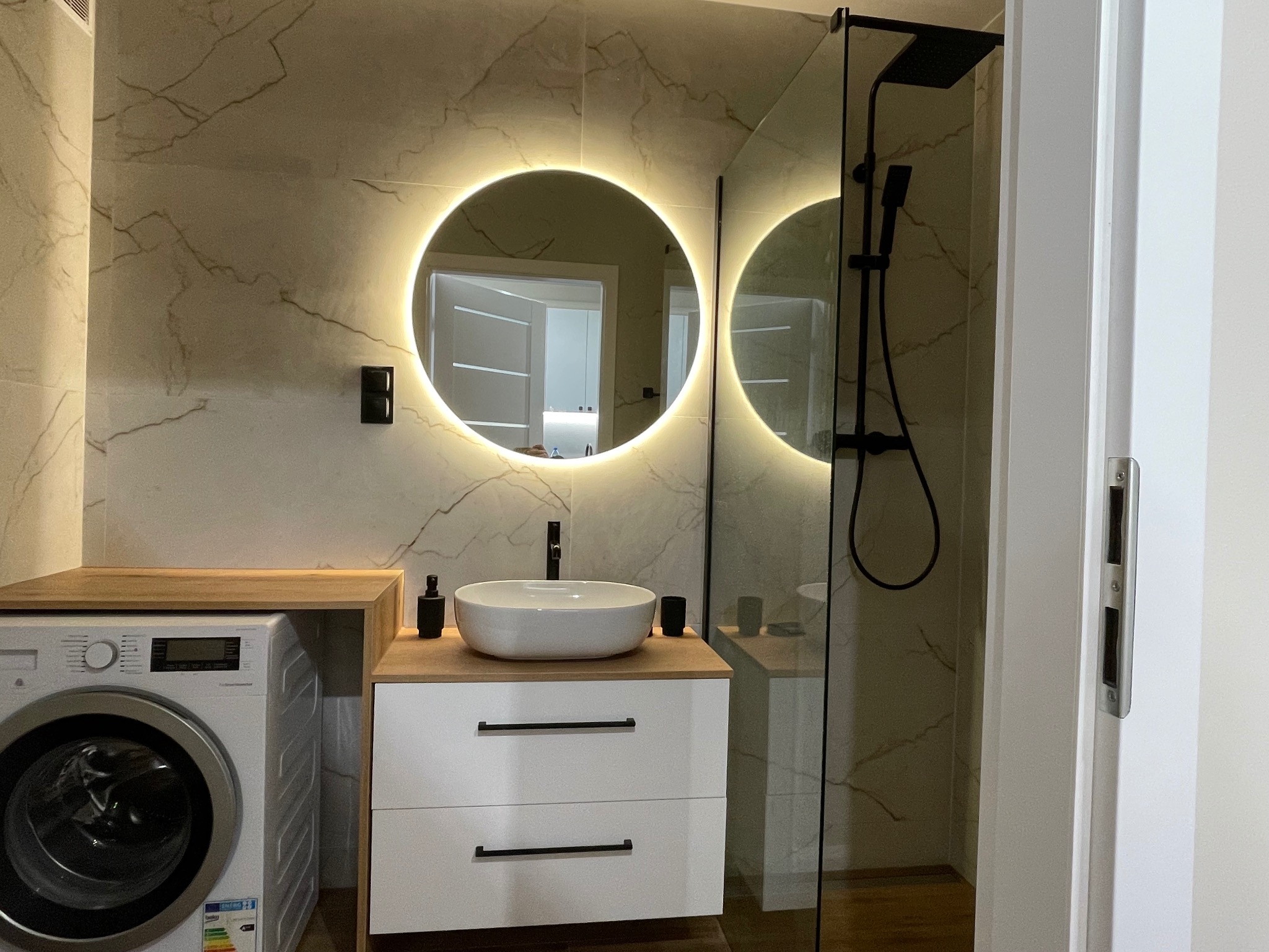 Nowoczesna łazienka z okrągłym lustrem LED, białą umywalką nablatową, pralką pod blatem i kabiną prysznicową typu walk-in z czarną armaturą na marmurowych płytkach.