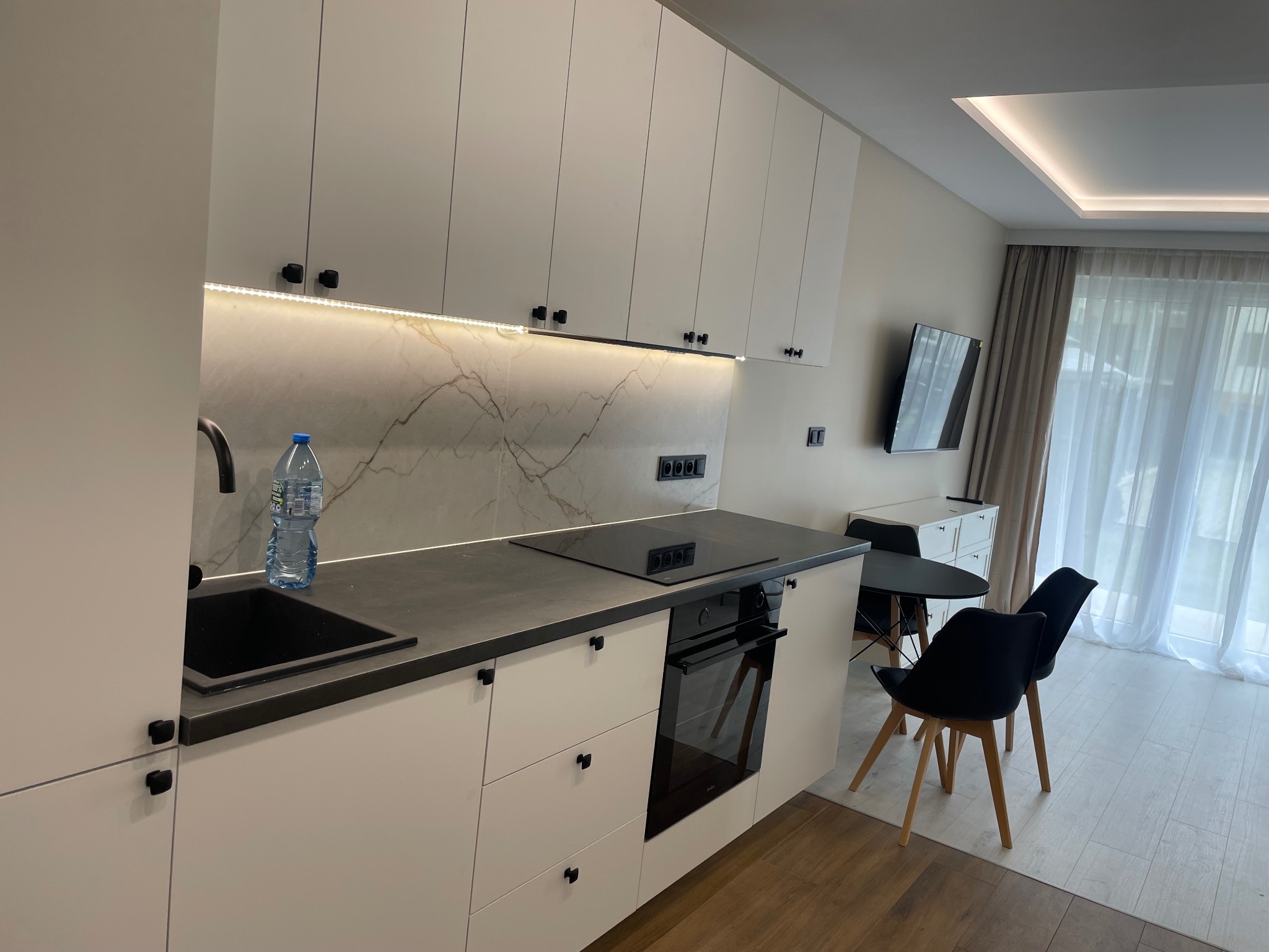 Nowoczesna, jasna kuchnia z białymi szafkami, czarnym blatem i marmurowym panelem. Oświetlenie LED pod szafkami. Widoczny zlew, płyta grzewcza i piekarnik.