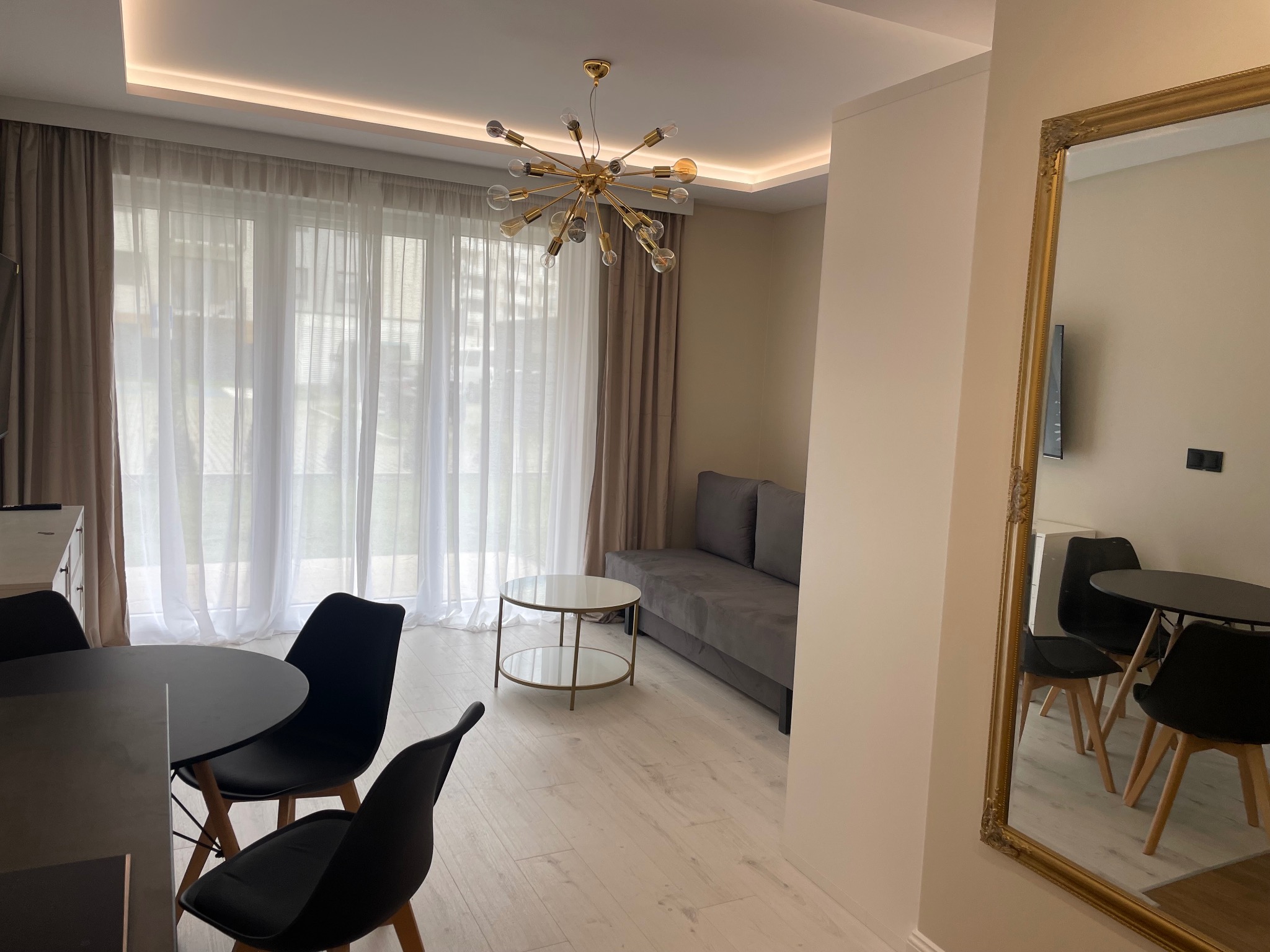 Jasne wnętrze salonu z parkietem: złoty żyrandol, szara sofa, okrągły stolik kawowy i stół z krzesłami, odbicie w lustrze w złotej ramie. Subtelne zasłony w oknie.