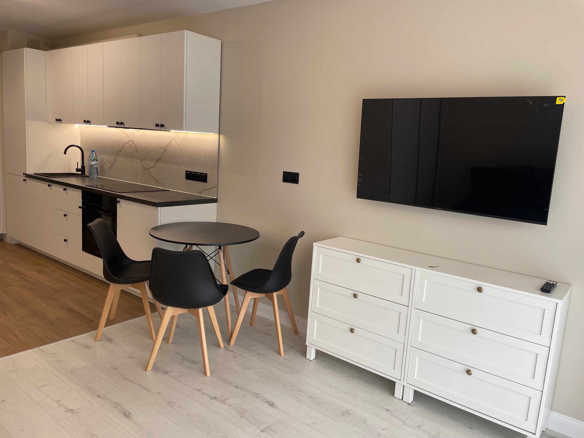 Nowoczesna aranżacja wnętrza: biała zabudowa kuchenna z marmurowym akcentem, czarne krzesła przy okrągłym stole i biała komoda pod telewizorem. Jasna podłoga.