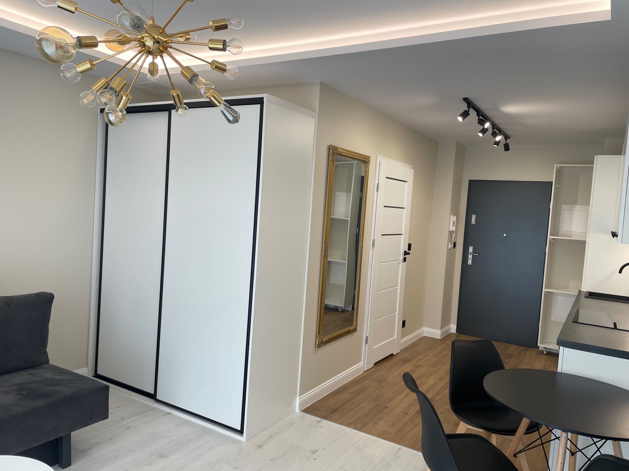 Nowoczesne wnętrze mieszkania: biała szafa w zabudowie, złoty żyrandol, lustro w zdobionej ramie i minimalistyczne meble w neutralnej kolorystyce.
