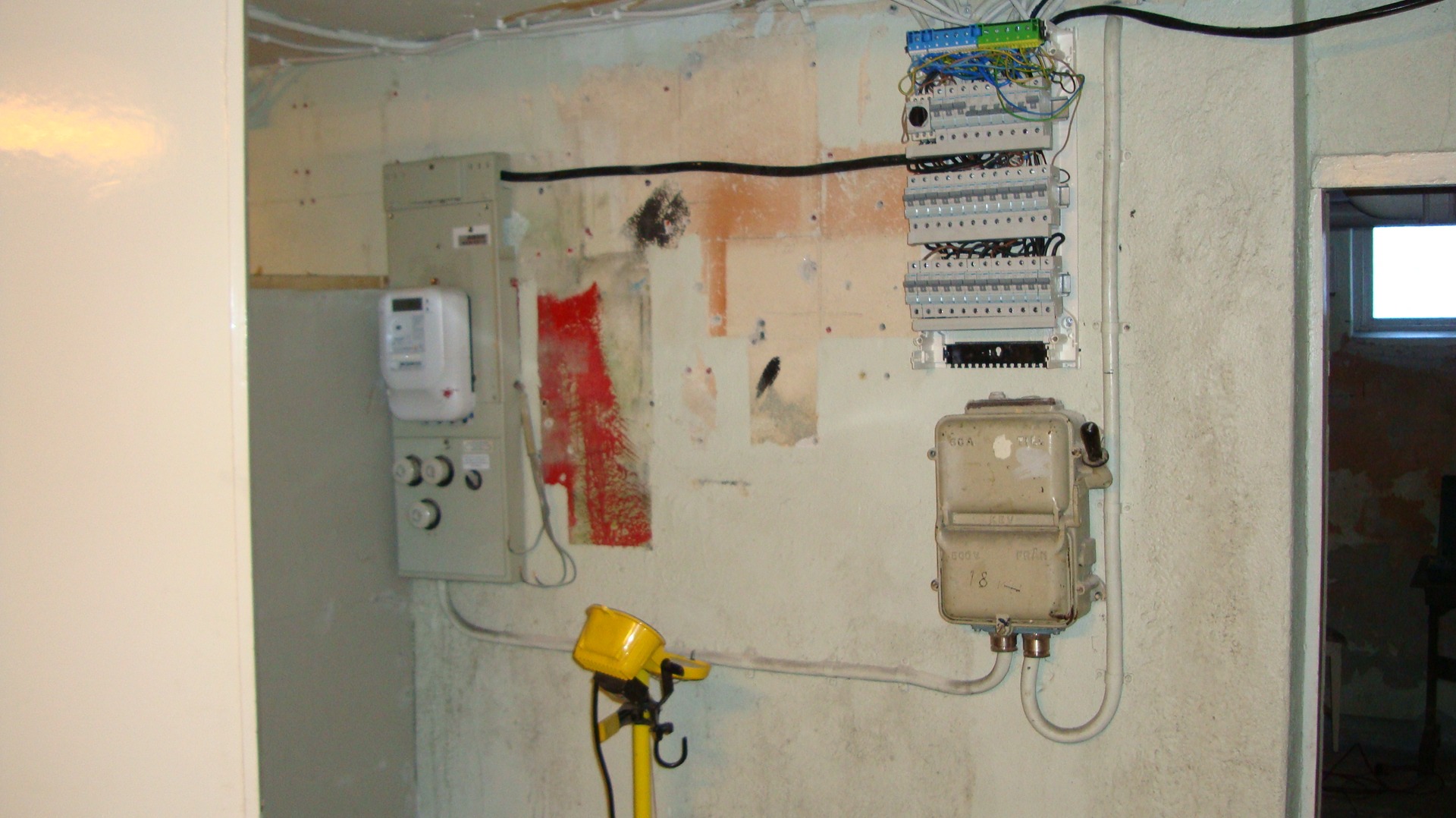 Rozdzielnica elektryczna ze starym licznikiem, zabezpieczeniami i przewodami na ścianie z widocznymi śladami po malowaniu, oświetlona żółtą lampą halogenową na statywie.