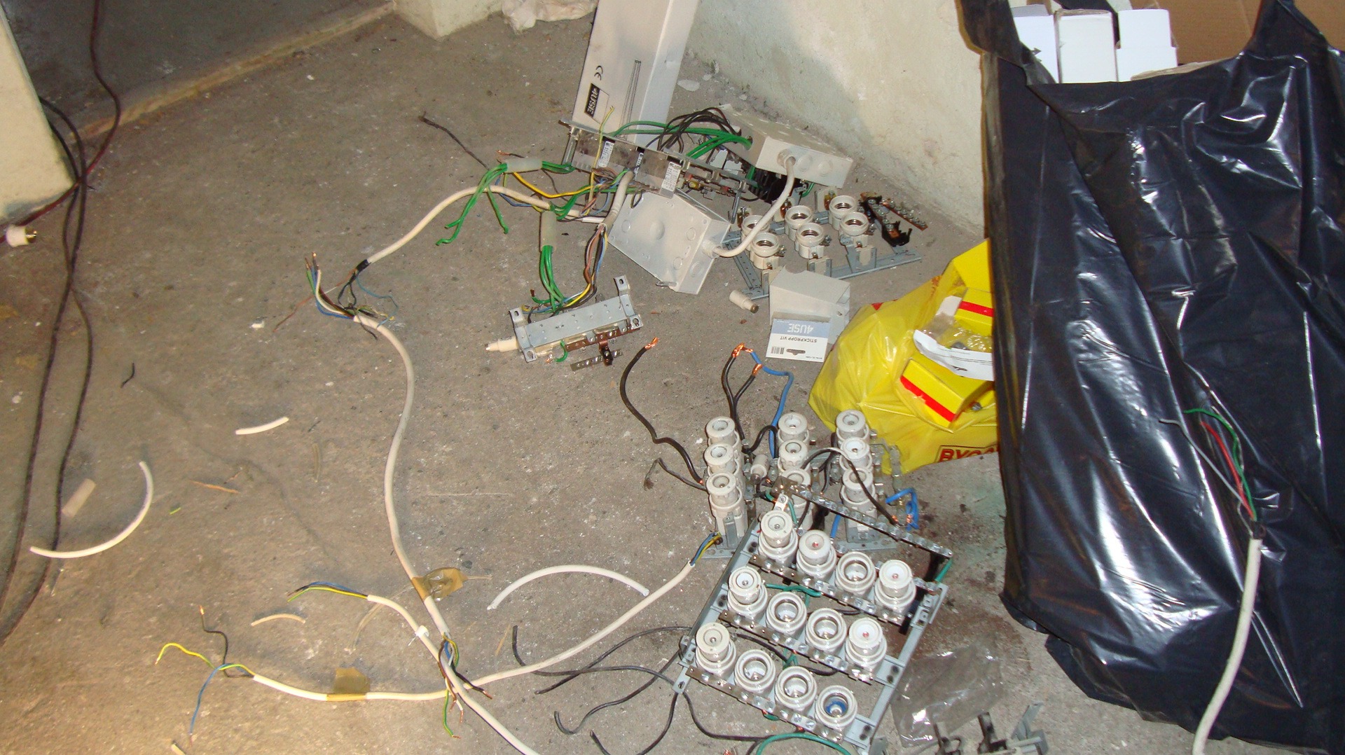 Rozmontowana skrzynka bezpiecznikowa z powypinanymi bezpiecznikami, przewodami i innymi elementami elektrycznymi leżąca na betonowej podłodze w nieładzie, obok czarny worek na śmieci.