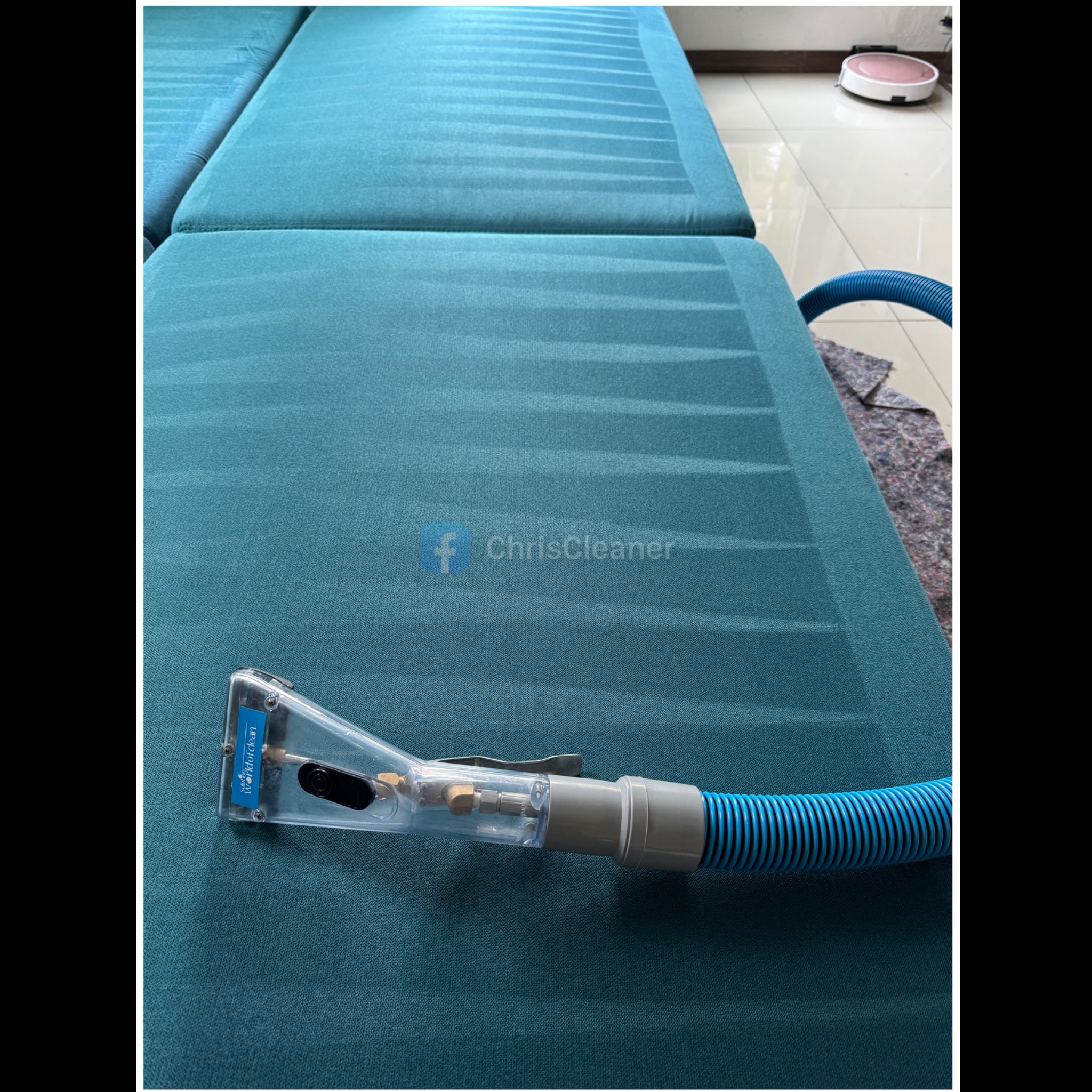 Turkusowa kanapa z widocznymi śladami czyszczenia, na której leży przezroczysta dysza odkurzacza piorącego z niebieskim wężem, w tle robot sprzątający.