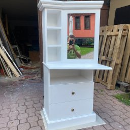 Leśnicki Stolarstwo - Biała, drewniana toaletka z lustrem, widoczne dwie szuflady z okrągłymi uchwytami, półki i odblask osoby robiącej zdjęcie telefonem, ustawiona na kostce brukowej, w tle warsztat i palety.