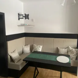 Wnęka jadalniana z tapicerowanym siedziskiem w kolorze beżowym, czarnym stołem z zielonym bieżnikiem i okrągłą podkładką, uzupełniona dekoracyjnymi poduszkami we wzory.