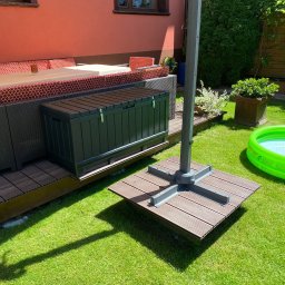 Leśnicki Stolarstwo - Zamykana skrzynia ogrodowa z tworzywa sztucznego w kolorze grafitowym, umieszczona na tarasie z desek kompozytowych, obok podstawy parasola przeciwsłonecznego i dmuchanego basenu.