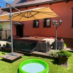 Leśnicki Stolarstwo - Zadaszony taras z drewnianą podłogą, meblami ogrodowymi, oświetleniem i basenem dla dzieci na trawniku przed domem.