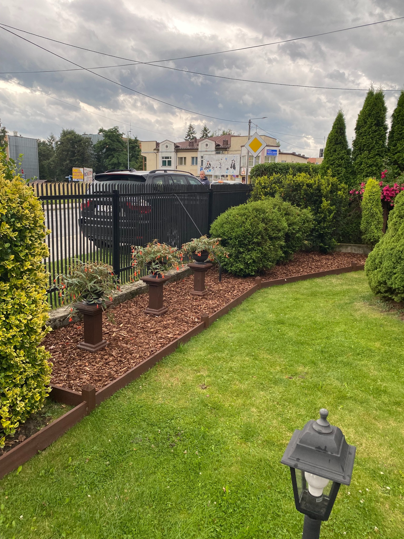 Uporządkowany ogród z rabatą z kory, obrzeżem z tworzywa imitującego drewno, roślinami w donicach na postumentach i czarnym ogrodzeniem panelowym w tle.