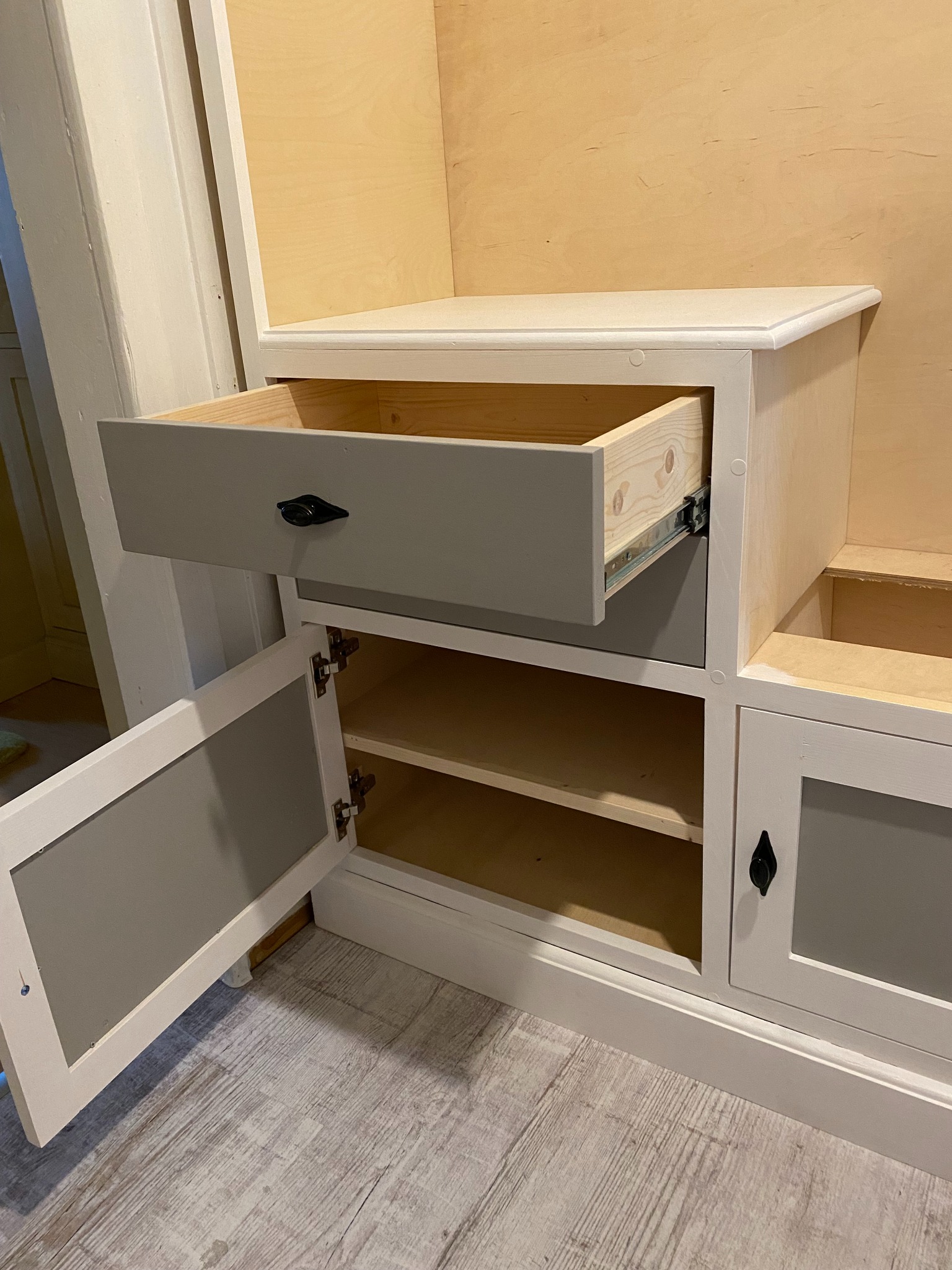 Biało-szara zabudowa meblowa z otwartą szufladą na metalowych prowadnicach i uchylonymi drzwiczkami, widoczne półki wewnątrz.