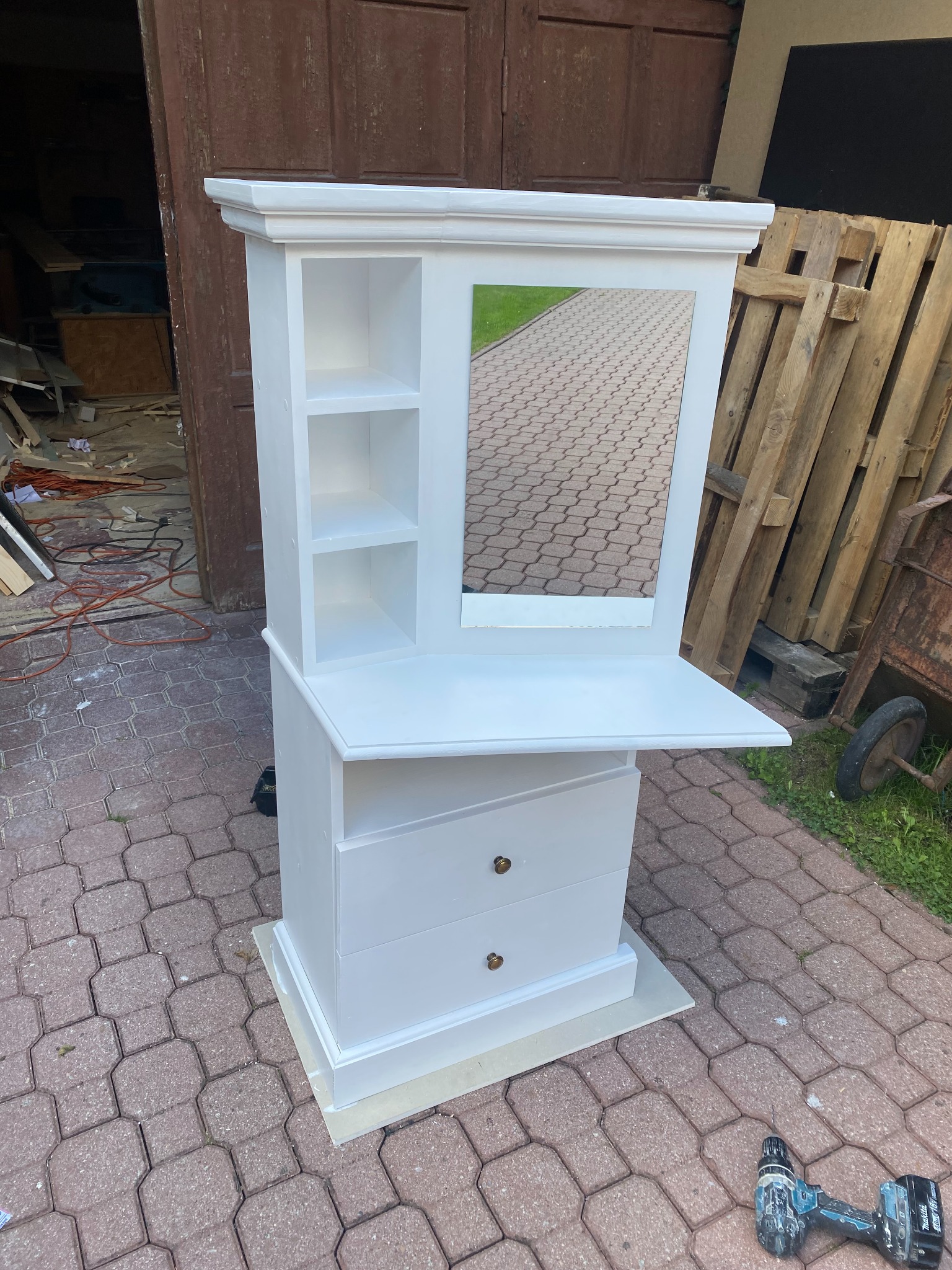 Biała toaletka z lustrem i wysuwanym blatem, widoczna na zewnątrz na tle palet i narzędzi, sugerująca etap wykończeniowy.