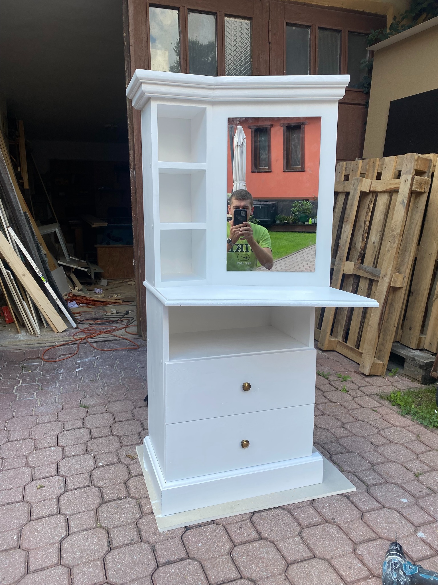 Biała, drewniana toaletka z lustrem, widoczne dwie szuflady z okrągłymi uchwytami, półki i odblask osoby robiącej zdjęcie telefonem, ustawiona na kostce brukowej, w tle warsztat i palety.