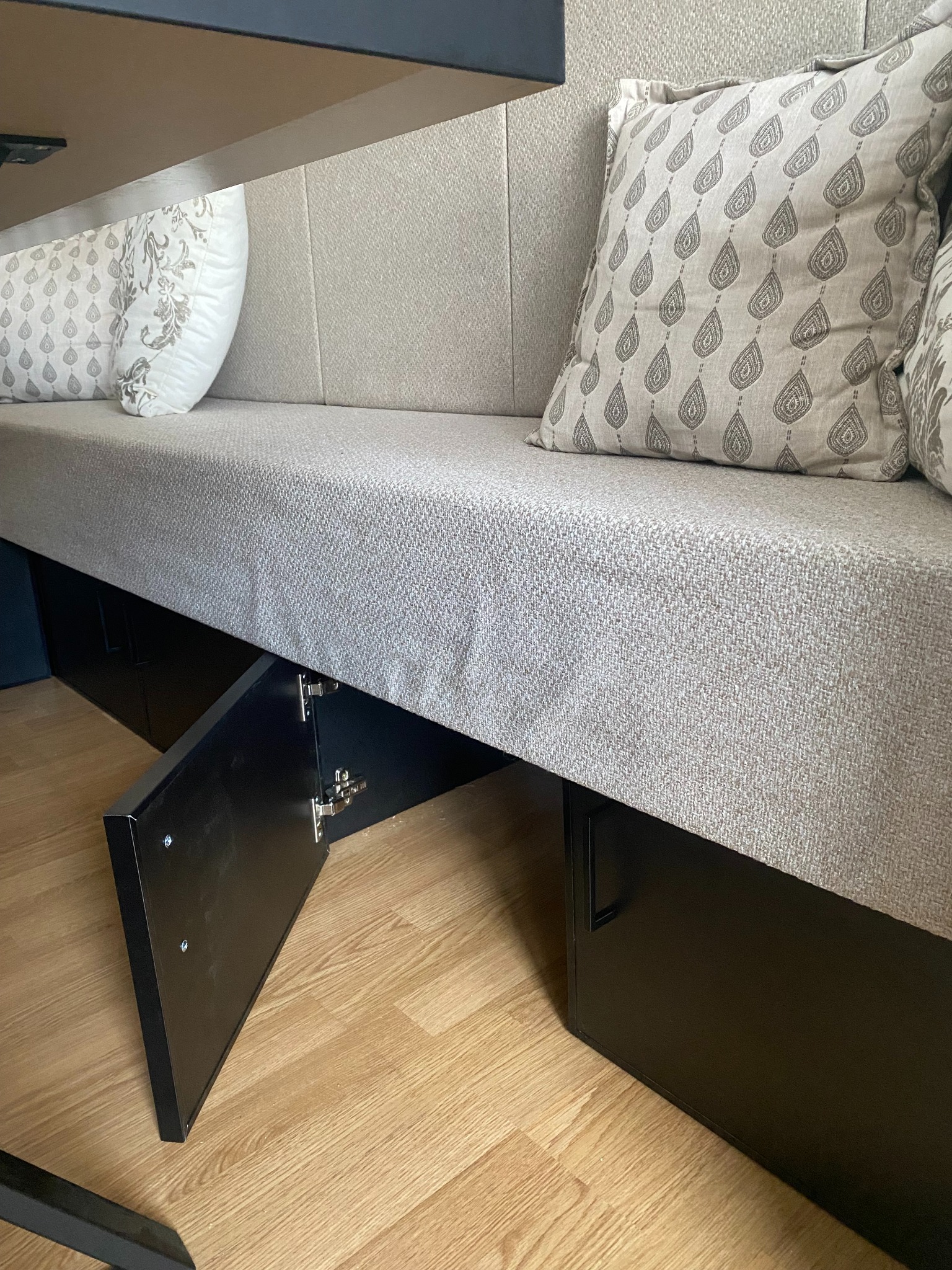 Tapicerowana ławka z podnoszonym siedziskiem, odsłaniającym schowek z czarnymi drzwiczkami na zawiasach, uzupełniona o dwie poduszki dekoracyjne.