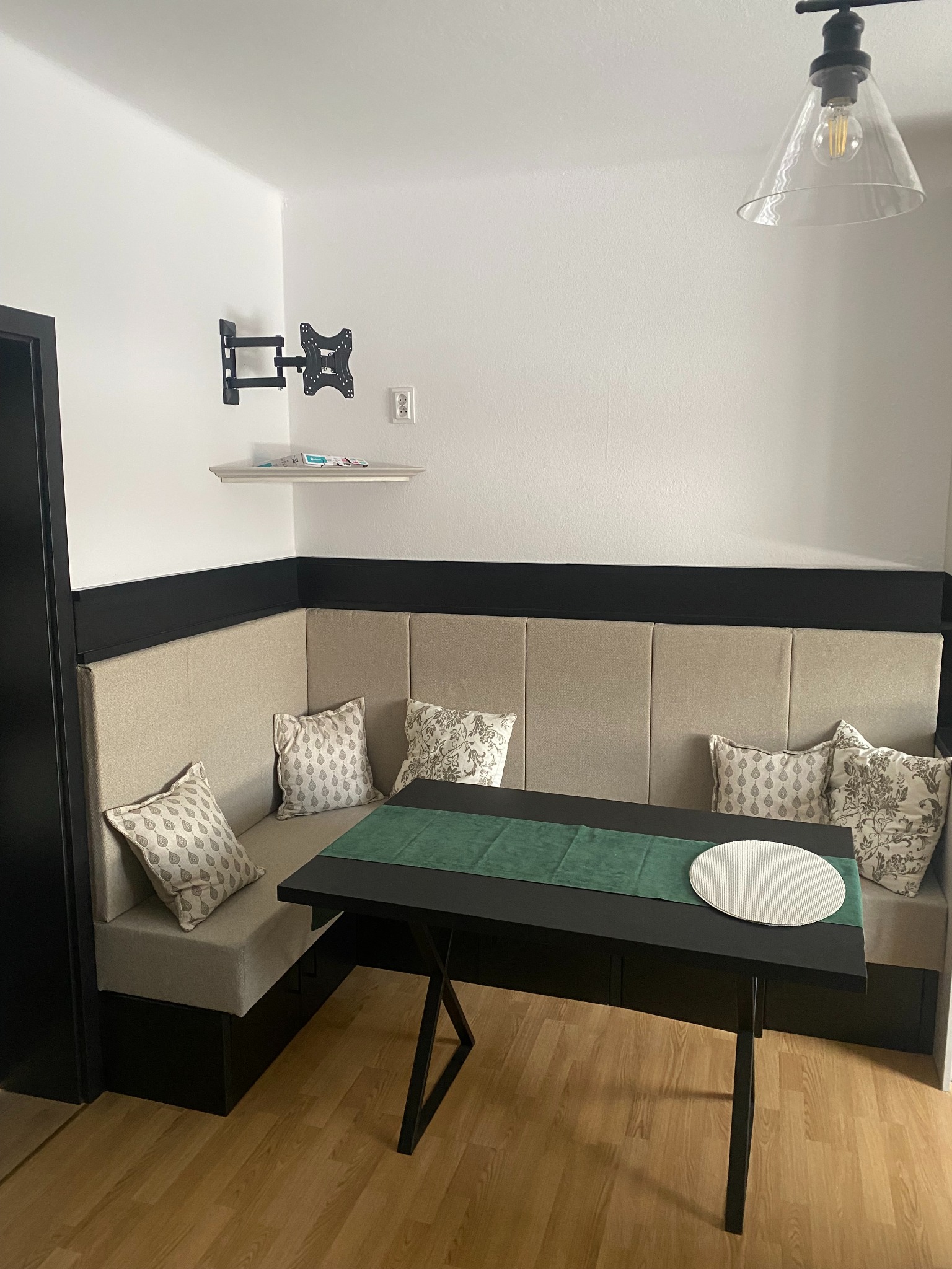 Wnęka jadalniana z tapicerowanym siedziskiem w kolorze beżowym, czarnym stołem z zielonym bieżnikiem i okrągłą podkładką, uzupełniona dekoracyjnymi poduszkami we wzory.