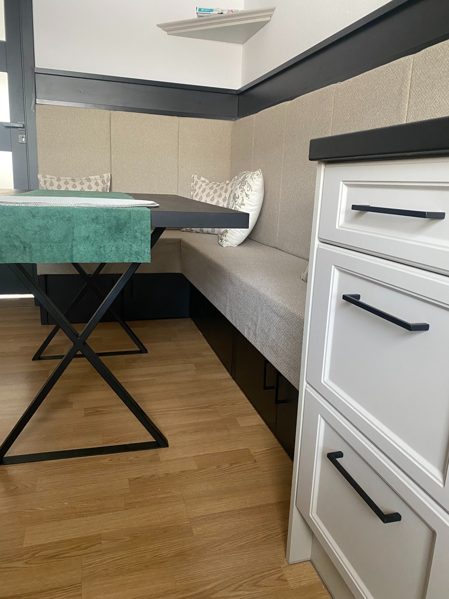 Wnętrze jadalni z narożną zabudową tapicerowaną, stół z czarnym blatem i metalowymi nogami, obok biała zabudowa meblowa z czarnymi uchwytami, podłoga drewniana.