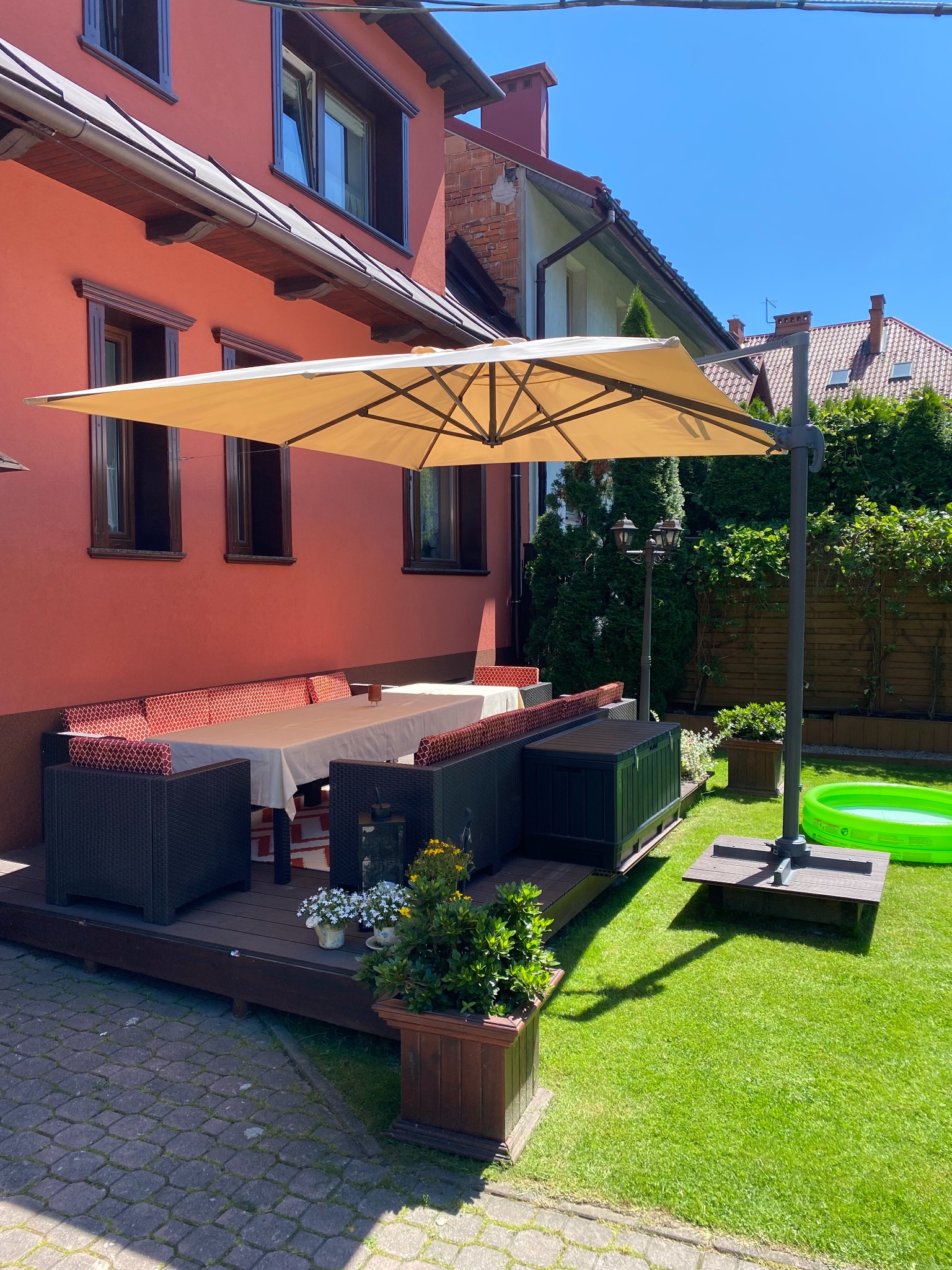 Zadaszony taras drewniany z meblami ogrodowymi i parasolem, obok zielony trawnik z basenem dla dzieci.