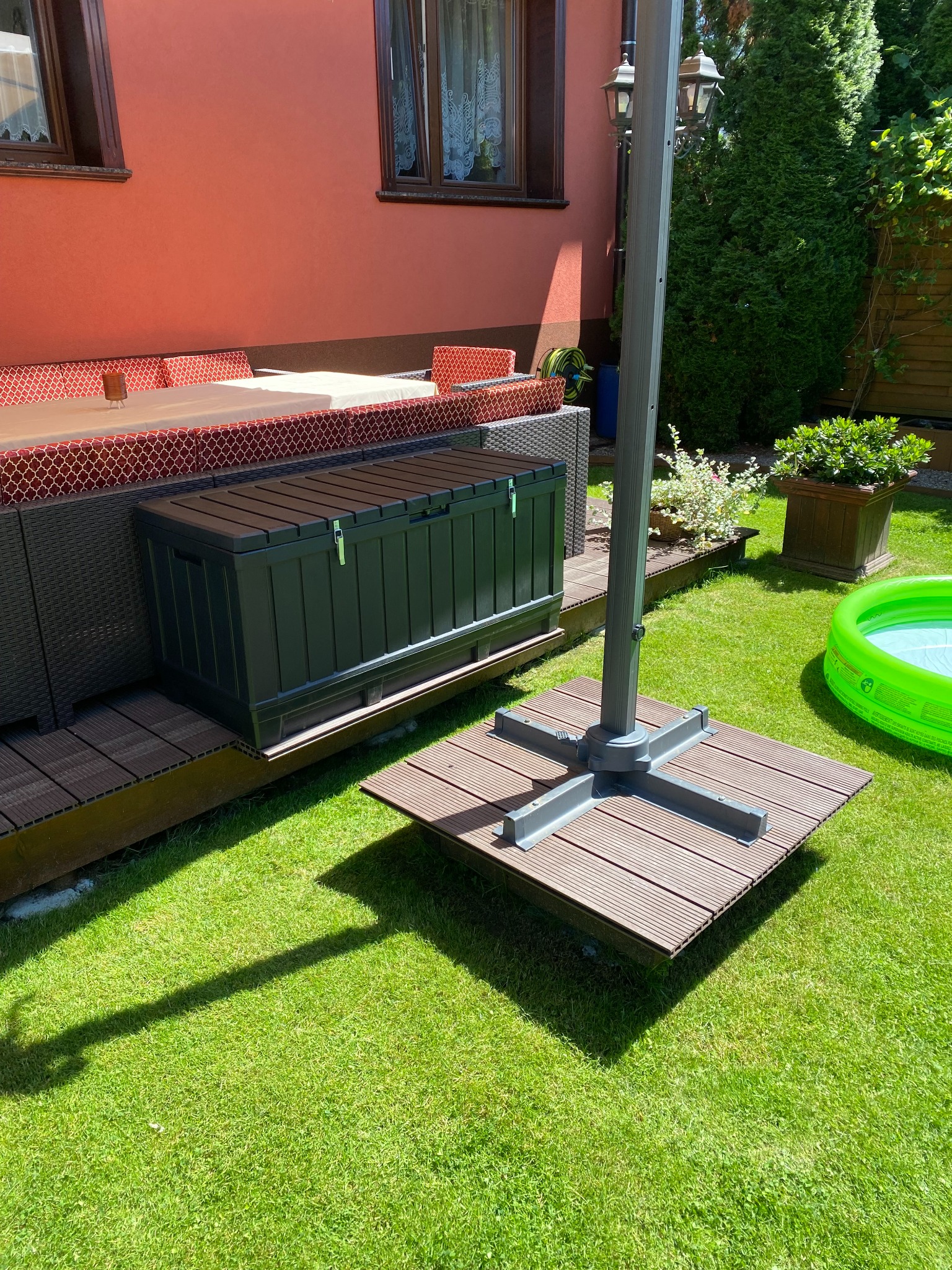 Zamykana skrzynia ogrodowa z tworzywa sztucznego w kolorze grafitowym, umieszczona na tarasie z desek kompozytowych, obok podstawy parasola przeciwsłonecznego i dmuchanego basenu.