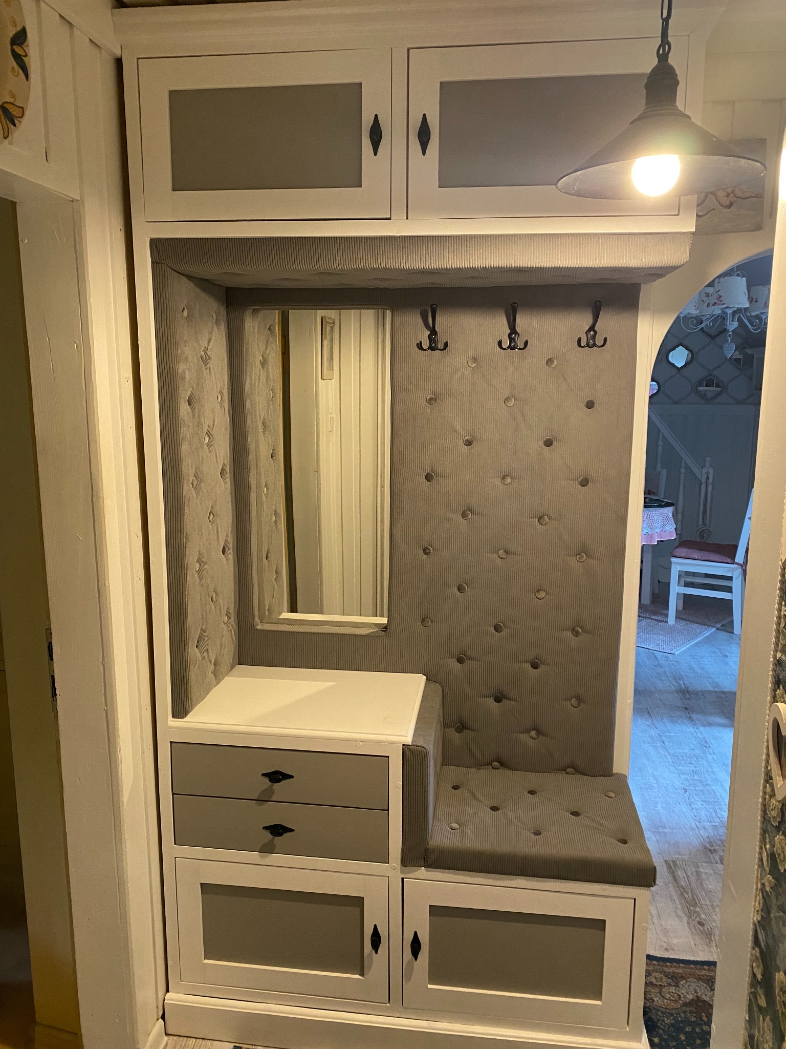 Wnęka z siedziskiem i lustrem w przedpokoju, obita szarym materiałem z guzikami, nad nią szafki i wieszaki, oświetlona wiszącą lampą.