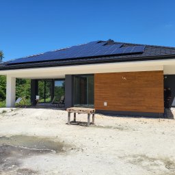 TERMIKA Paweł Józwik - Nowoczesny dom parterowy z panelami słonecznymi na dachu. Elewacja z drewnianą okładziną i dużymi przeszkleniami. Teren wokół domu w trakcie prac wykończeniowych.