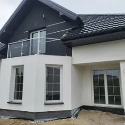 Nowoczesny dom jednorodzinny z białą elewacją i czarnym dachem, balkon z szklaną balustradą. Widoczne okna i fragment terenu wokół budynku.