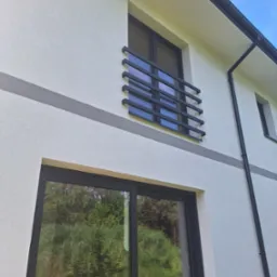 Biała elewacja domu z szarym pasem, czarnymi oknami i balkonem. Widok z dołu ukazuje fakturę tynku i detale architektoniczne. Błękitne niebo w tle.