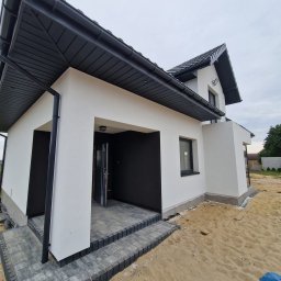 TERMIKA Paweł Józwik - Elewacja domu jednorodzinnego po malowaniu w Lubochni. Białe ściany, czarne elementy dachu i stolarki okiennej. Nowoczesny styl, kostka brukowa przed wejściem.