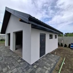 TERMIKA Paweł Józwik - Biały budynek gospodarczy z ciemnym dachem i drzwiami, świeżo po tynkowaniu. Widoczne detale wykończenia i kostka brukowa wokół budynku.