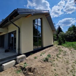 TERMIKA Paweł Józwik - Jasna elewacja domu z dużym oknem i szarą stolarką, widok z zewnątrz w słoneczny dzień. Elementy wykończeniowe przy dachu. Teren wokół domu w trakcie prac ziemnych.