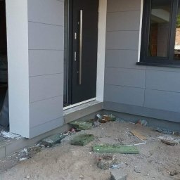 TERMIKA Paweł Józwik - Wykończona elewacja domu z szarymi panelami, czarnymi drzwiami i oknem. Widoczne pozostałości po pracach budowlanych na gruncie przed budynkiem. Ujęcie z bliska.