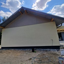 TERMIKA Paweł Józwik - Elewacja domu po tynkowaniu, z widoczną strukturą tynku i pomalowanym cokołem. W tle błękitne niebo z chmurami, na pierwszym planie wąż i wiadro po farbie.