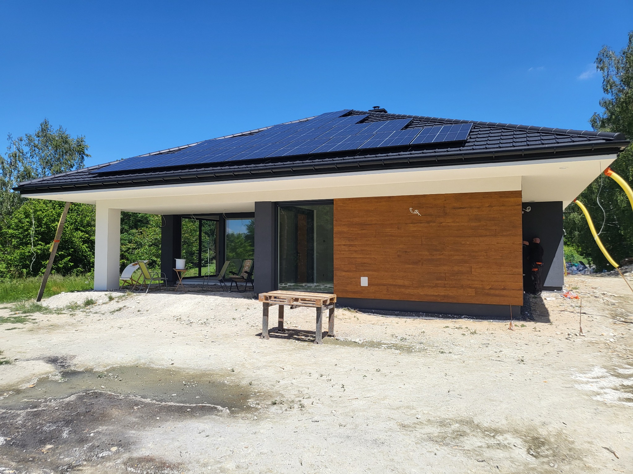 Nowoczesny dom parterowy z panelami słonecznymi na dachu. Elewacja z drewnianą okładziną i dużymi przeszkleniami. Teren wokół domu w trakcie prac wykończeniowych.