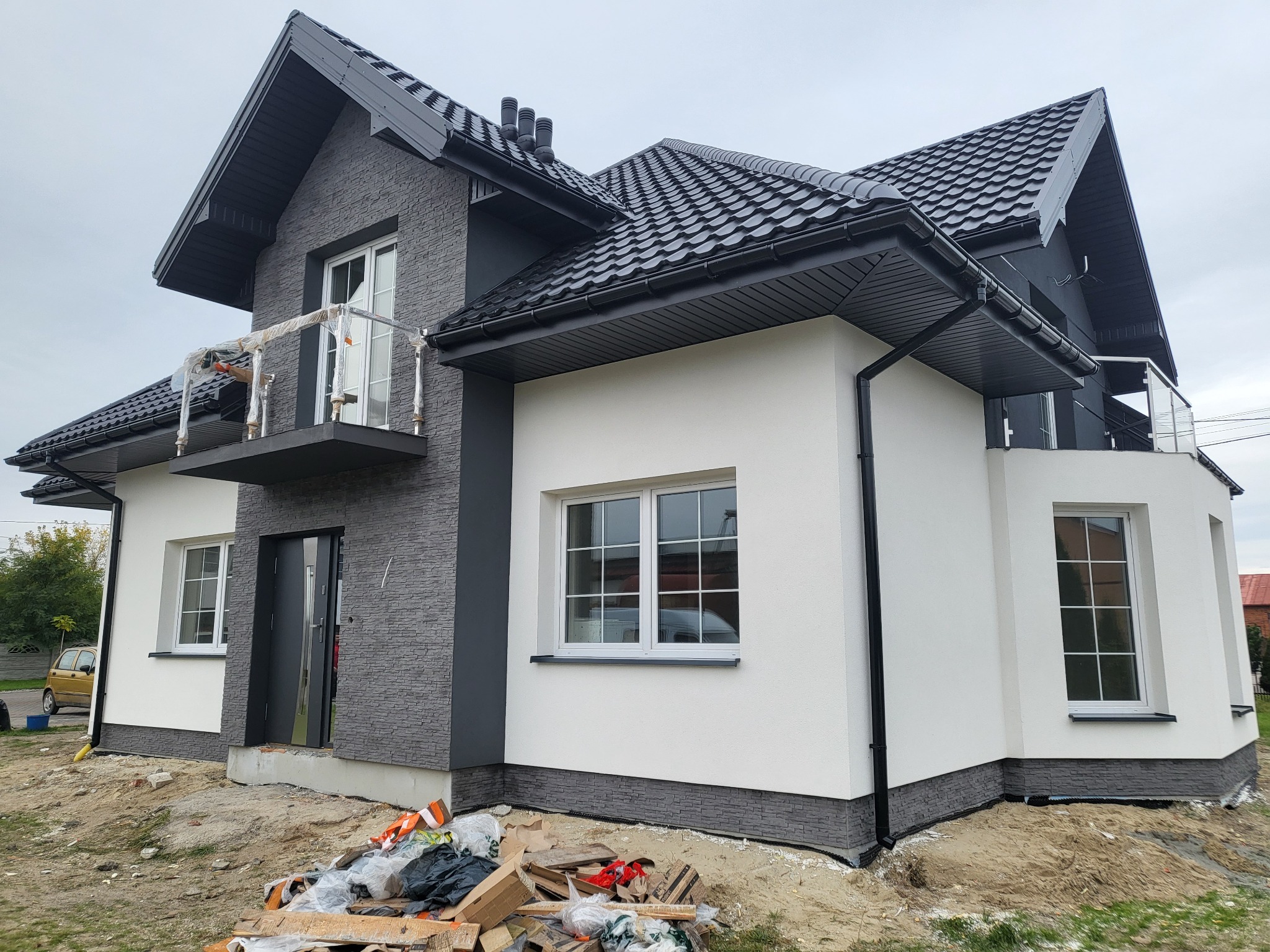 Nowoczesny dom z elewacją w odcieniach szarości i bieli, z balkonem i czarną dachówką. Widoczne prace wykończeniowe wokół budynku.